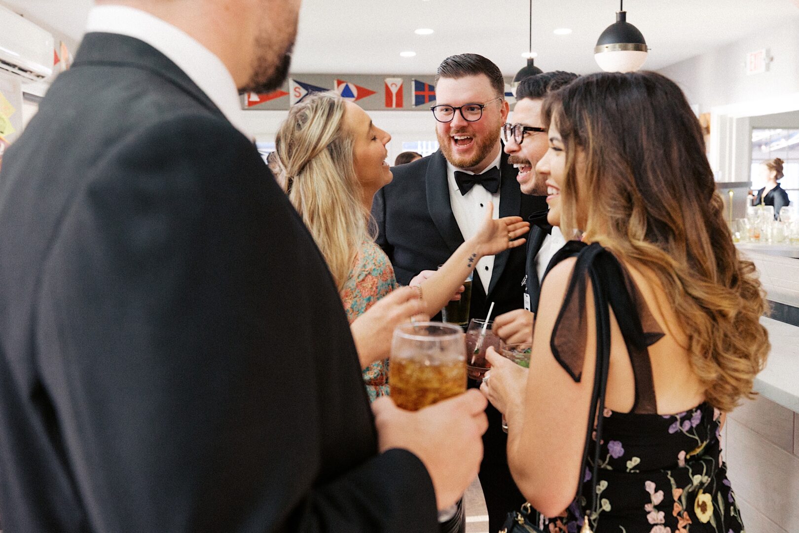 A group of people dressed formally are gathered indoors at the Corinthian Yacht Club of Cape May, smiling and holding drinks as they celebrate the joyful wedding occasion.