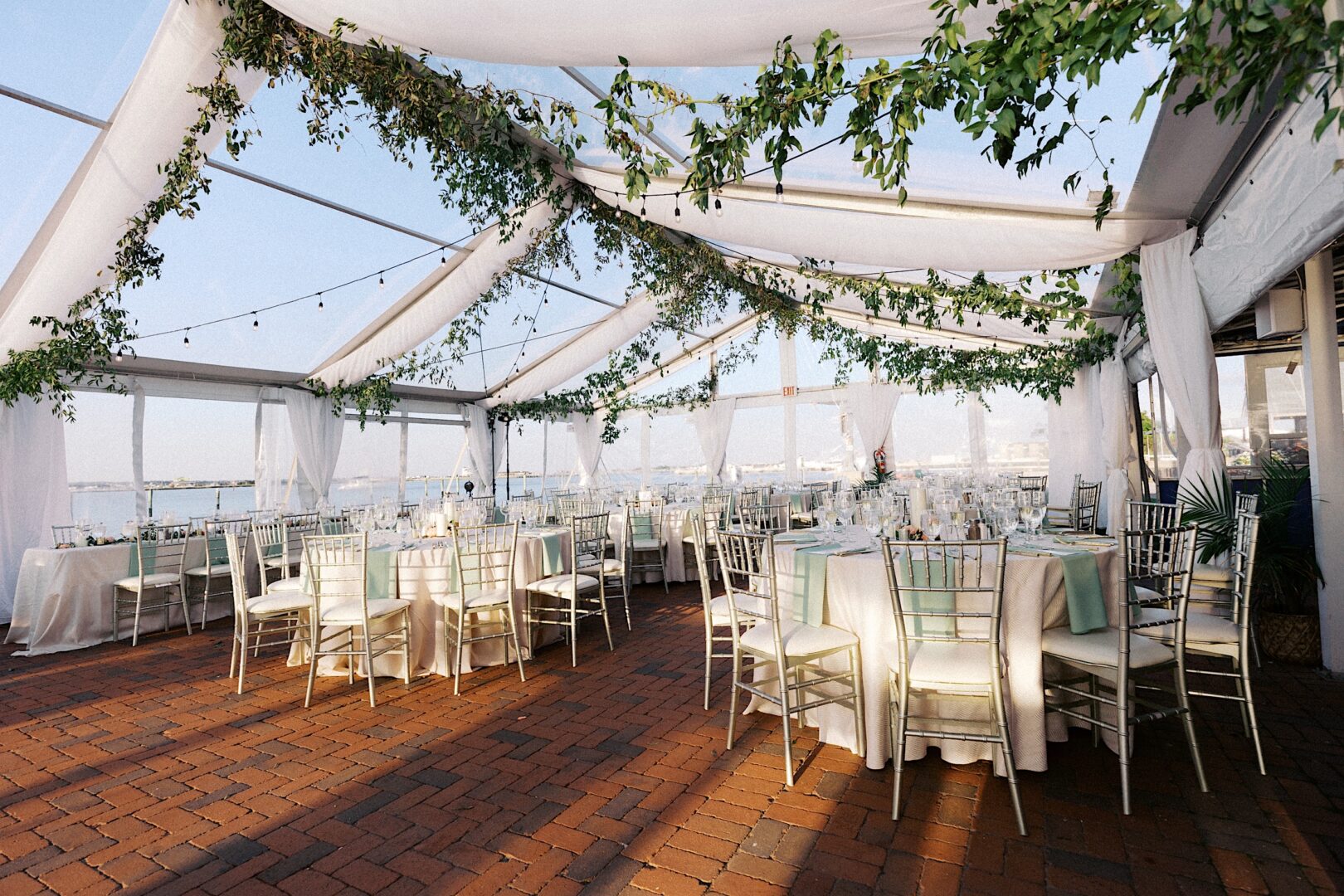 Outdoor wedding setup at the Corinthian Yacht Club of Cape May, featuring a transparent canopy with white draped fabric and lush greenery. Rows of white tables and chairs rest on the brick flooring, offering a stunning view of the water in the background.