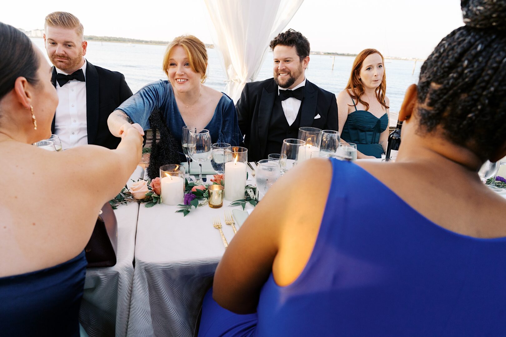 At a Corinthian Yacht Club of Cape May wedding, a group of elegantly dressed guests sits at a waterfront table. As the soothing candlelight flickers, one woman warmly shakes hands with another across the table, creating an atmosphere of sophistication and camaraderie.