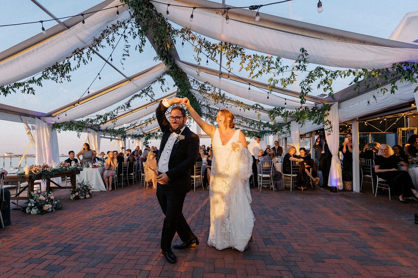 Under the airy embrace of a clear tent at the Corinthian Yacht Club of Cape May, a bride and groom share an enchanting dance. Lush greenery weaves through the decor, while their wedding guests look on in admiration.