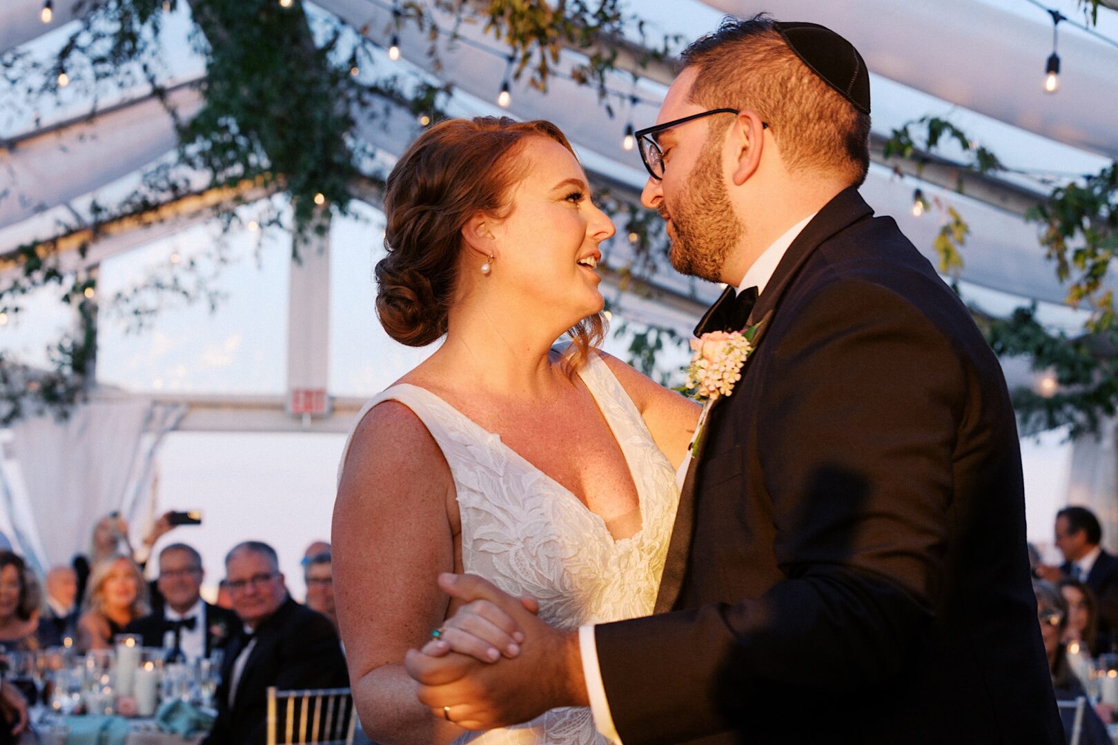 A bride and groom dance together under a tent at the Corinthian Yacht Club of Cape May wedding, with guests seated in the background. The ceiling is beautifully adorned with greenery and string lights. 