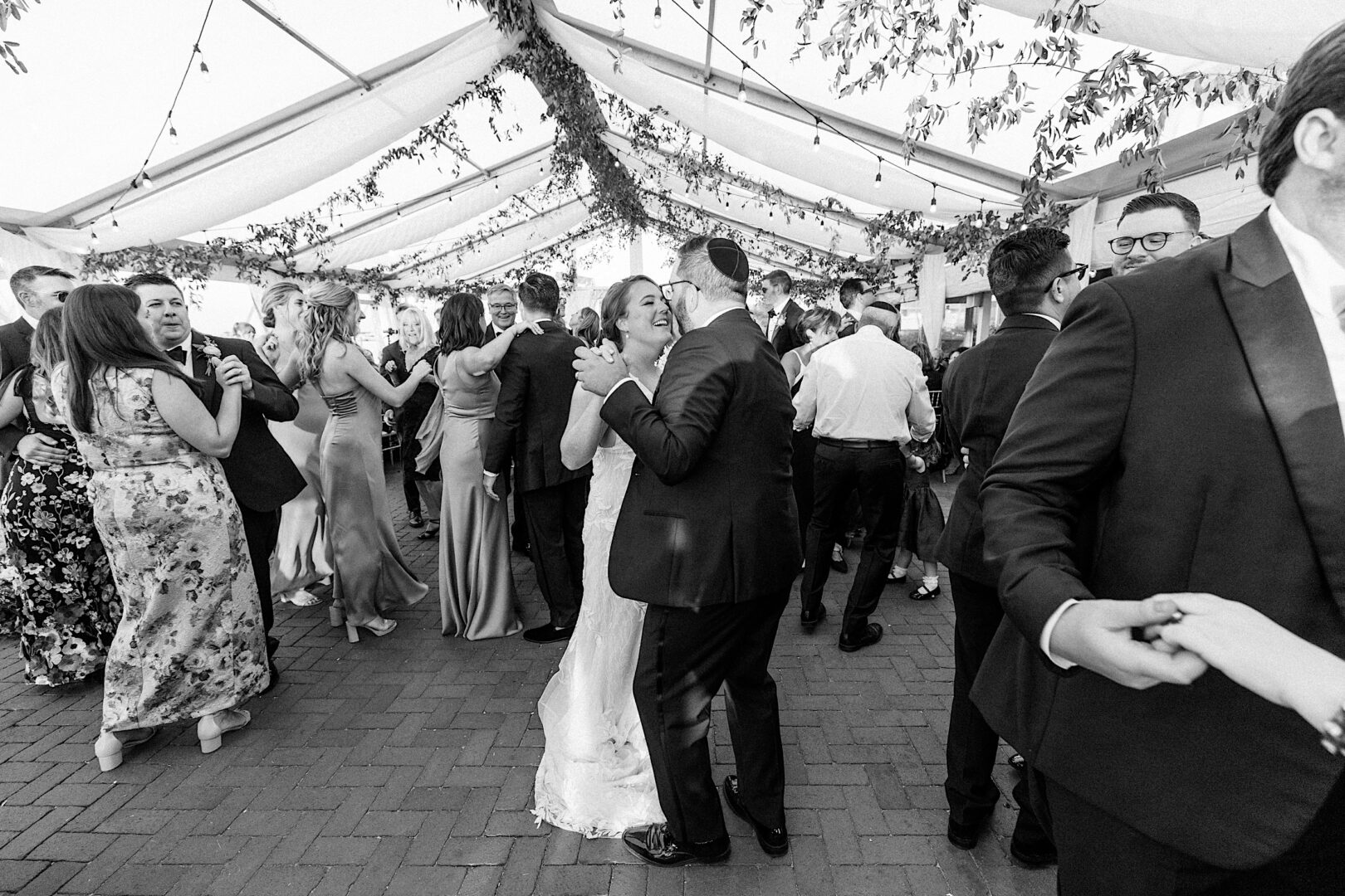 A Corinthian Yacht Club of Cape May wedding reception unfolds as couples dance under a tent adorned with greenery and string lights. Guests, draped in formal attire, celebrate the joyous occasion against the stunning waterfront backdrop.