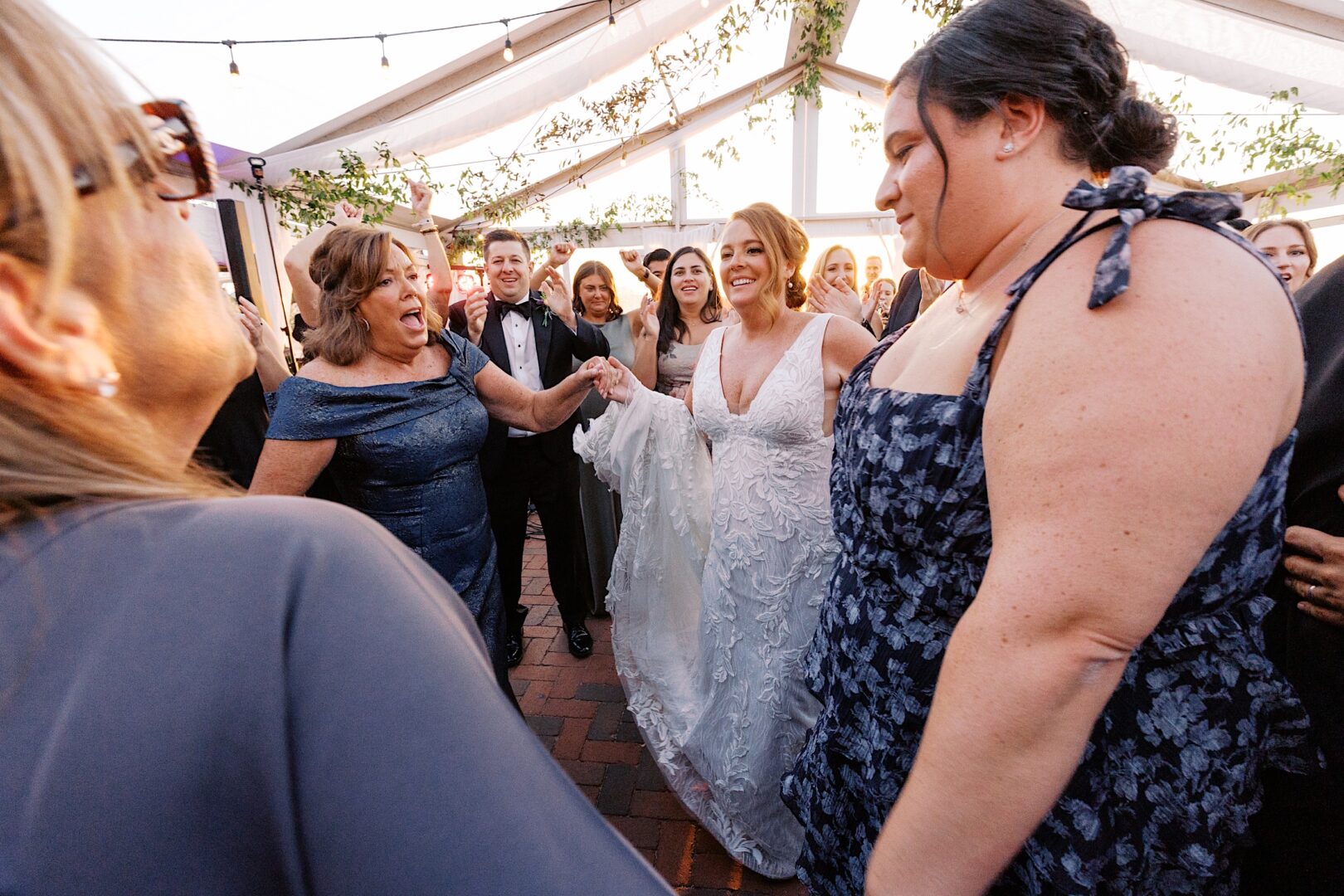 A group of people, including a bride in a white dress, are joyfully dancing together under a tent with string lights and greenery at the Corinthian Yacht Club of Cape May wedding.