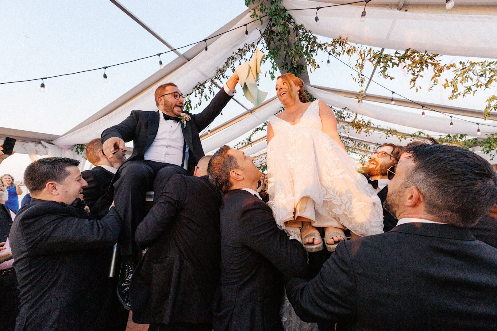 At the Corinthian Yacht Club of Cape May wedding, the bride and groom are joyously lifted on chairs by guests, surrounded by a tent beautifully adorned with vibrant greenery.