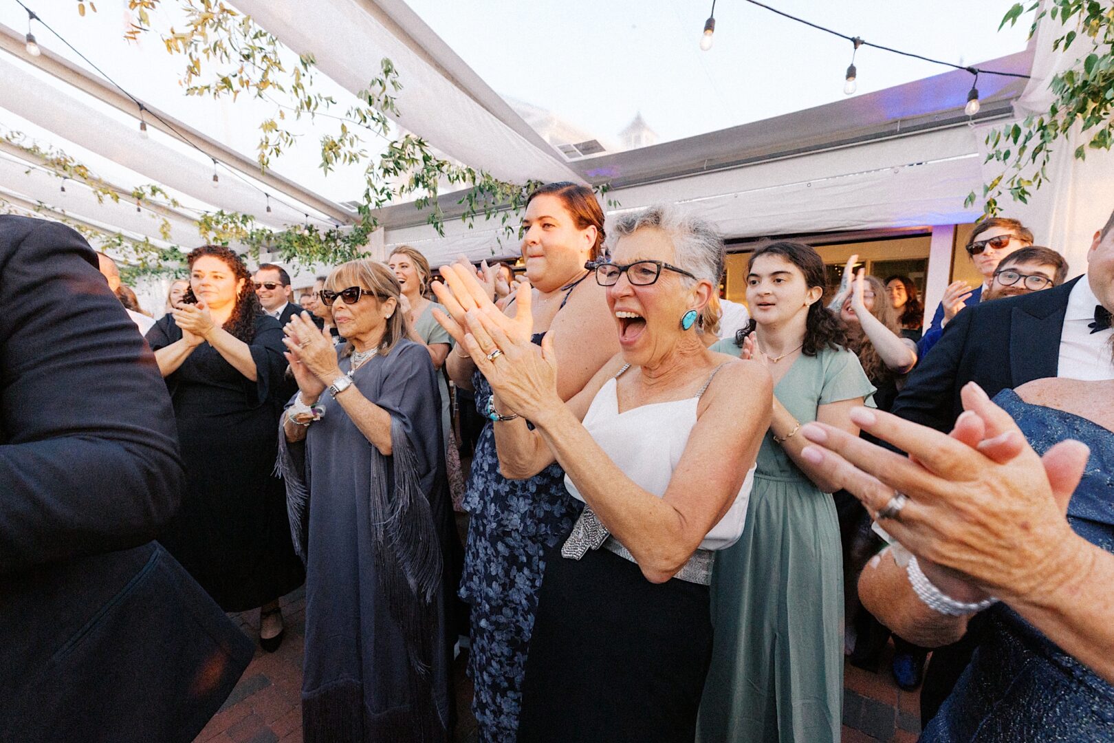 A group of people clapping and smiling in an outdoor setting under string lights and a white canopy at a picturesque Corinthian Yacht Club of Cape May wedding.