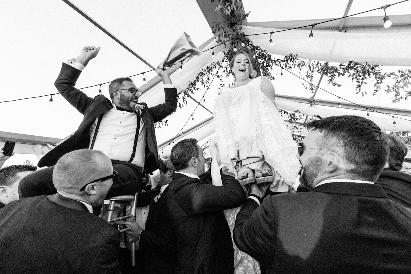 At the Corinthian Yacht Club of Cape May, a bride and groom are joyfully lifted on chairs by guests during a magical wedding celebration under a tent adorned with twinkling string lights.