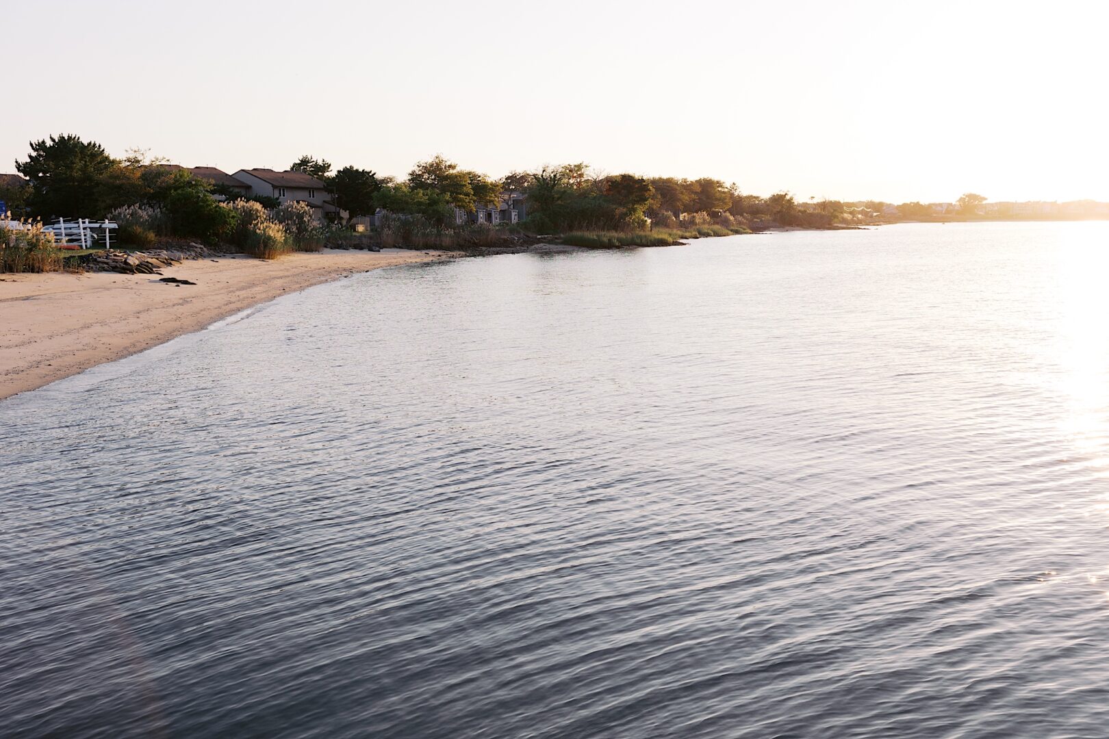 As the sun sets over a tranquil beach with gentle waves and a sandy shore, the Corinthian Yacht Club of Cape May Wedding graces the distant view, blending elegance with natural beauty. Houses in the distance add a charming touch to this picturesque scene.