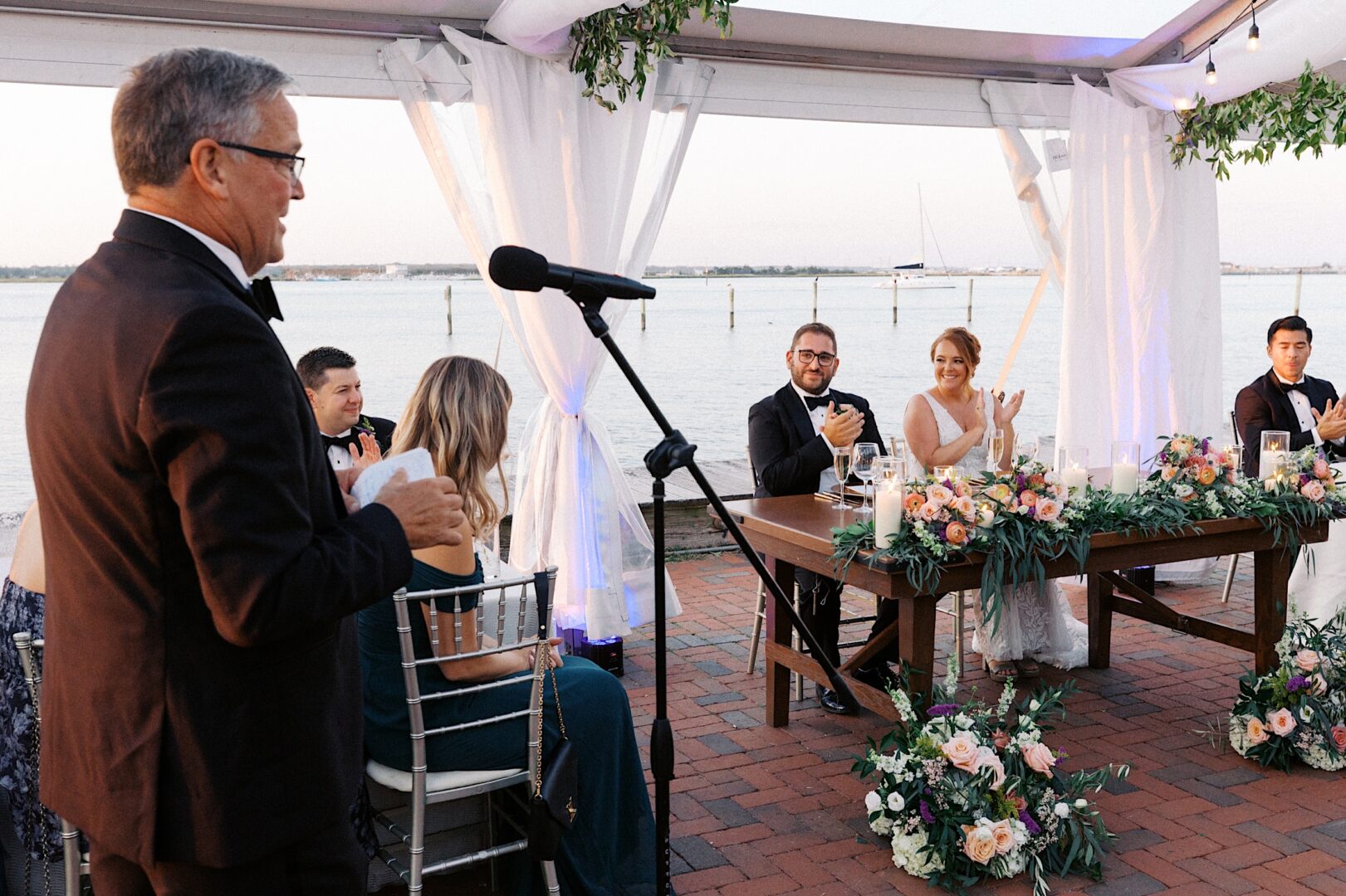 A man in a tuxedo speaks at a microphone during a wedding reception at the Corinthian Yacht Club of Cape May. Guests sit at tables near the water, surrounded by elegant floral arrangements and flickering candles.