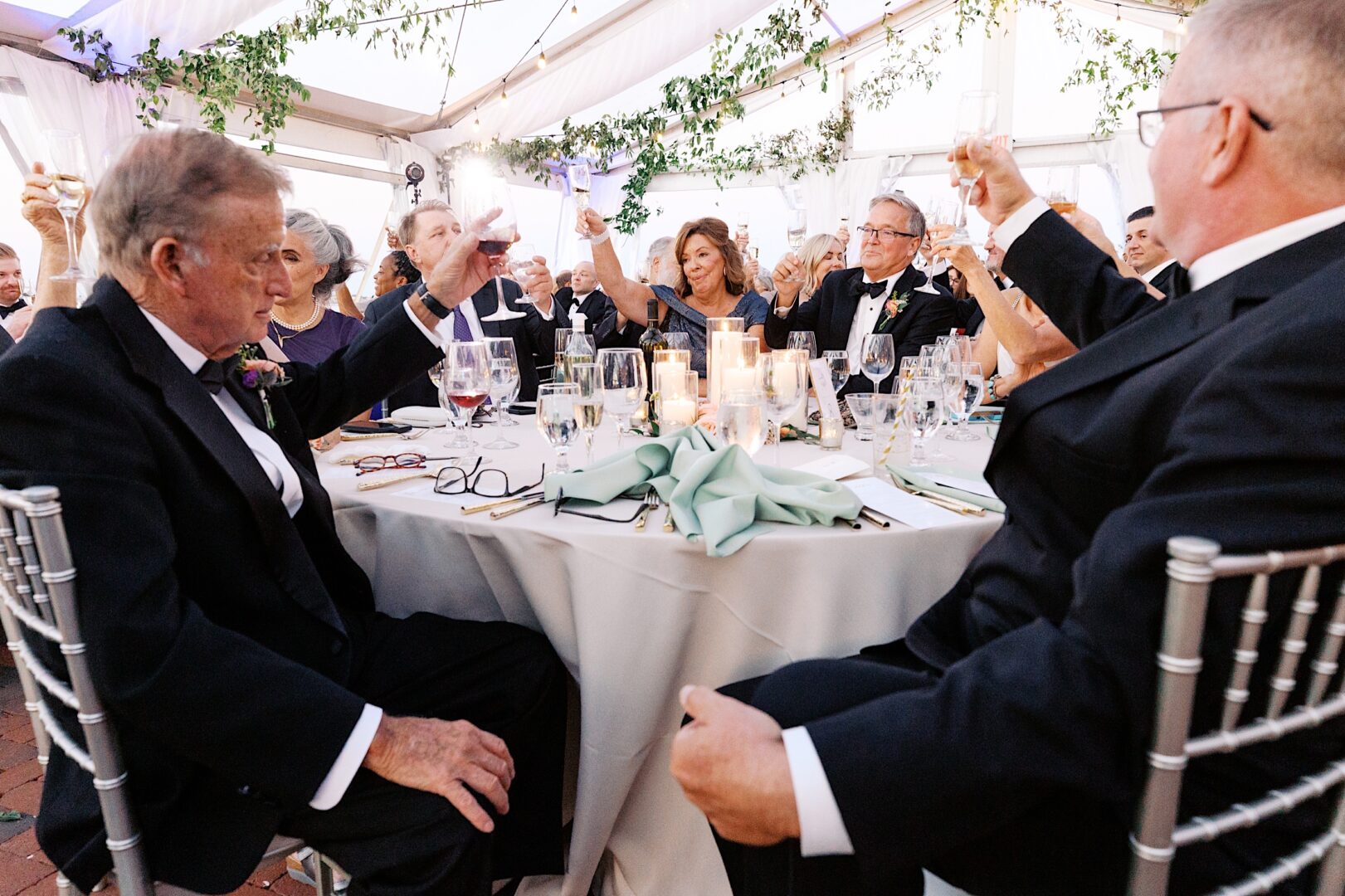 At the Corinthian Yacht Club of Cape May wedding, guests raise glasses for a toast while seated at a beautifully decorated table under an elegant tent.