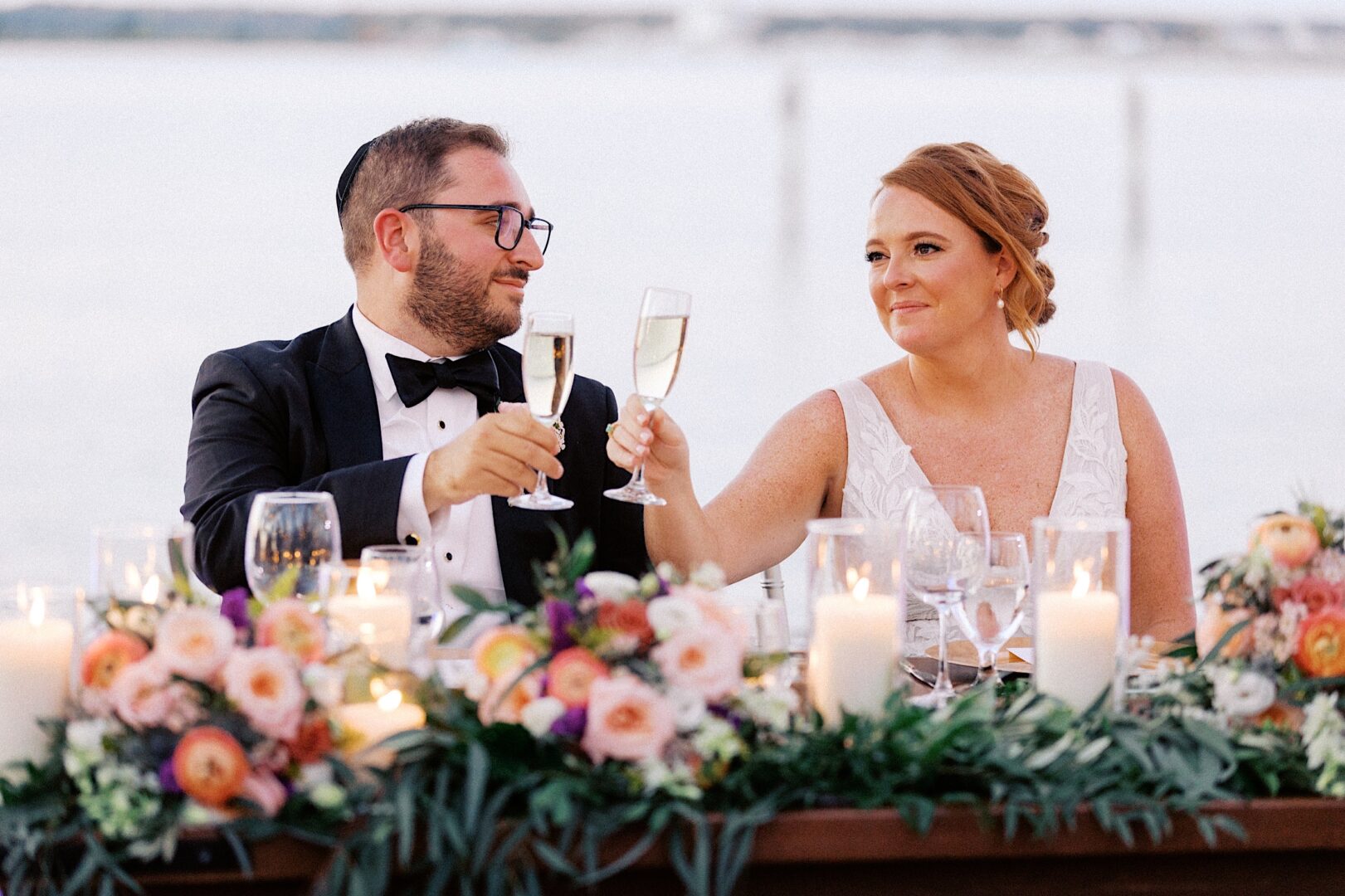 A couple in formal attire clinks champagne glasses at a decorated table adorned with candles and flowers, against the tranquil waterfront backdrop of the Corinthian Yacht Club of Cape May wedding.