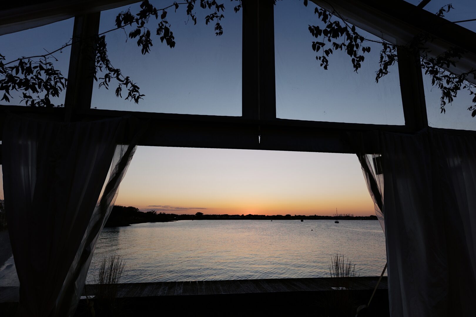 A serene view of a calm lake at sunset unfolds through the window with partially open curtains and silhouetted leaves above, evoking the tranquil elegance of a Corinthian Yacht Club of Cape May wedding.