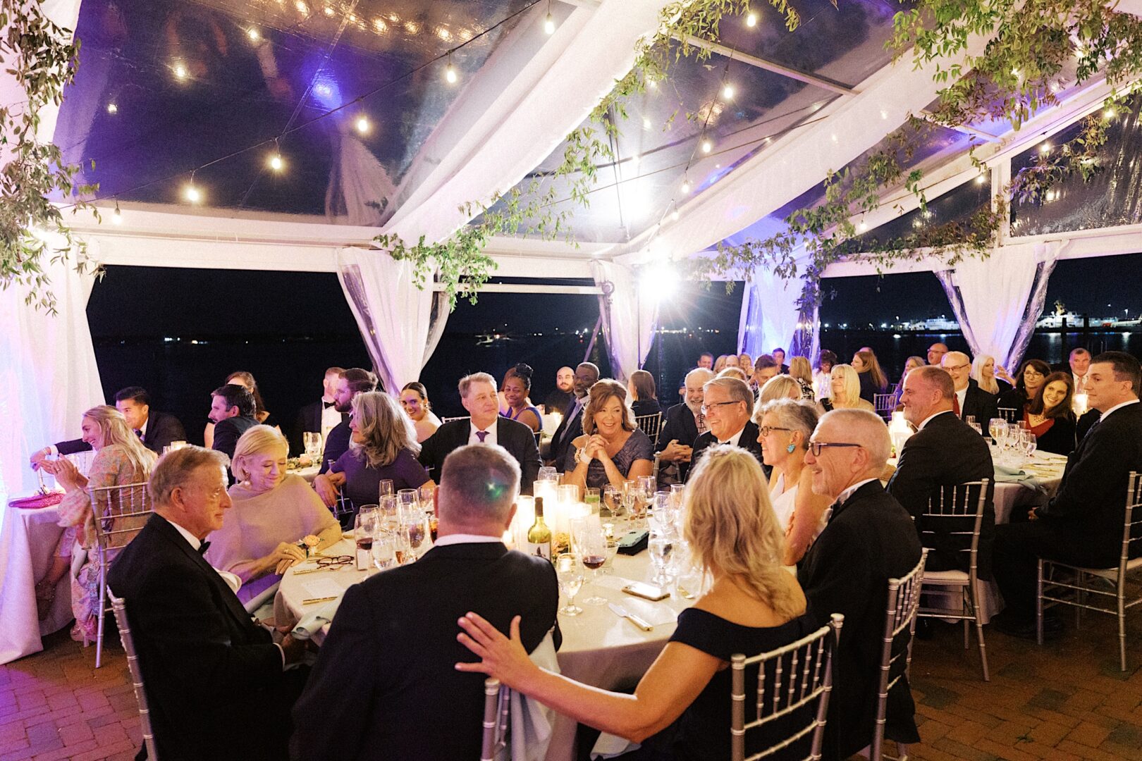 Guests seated at round tables under a decorated tent for an evening event, with string lights and greenery overhead, experience the charm of a Corinthian Yacht Club of Cape May wedding.