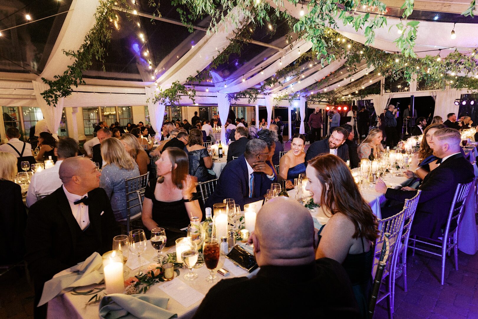 Guests in formal attire are seated at elegantly decorated tables under a canopy, with string lights and greenery creating a magical atmosphere at the Corinthian Yacht Club of Cape May wedding.