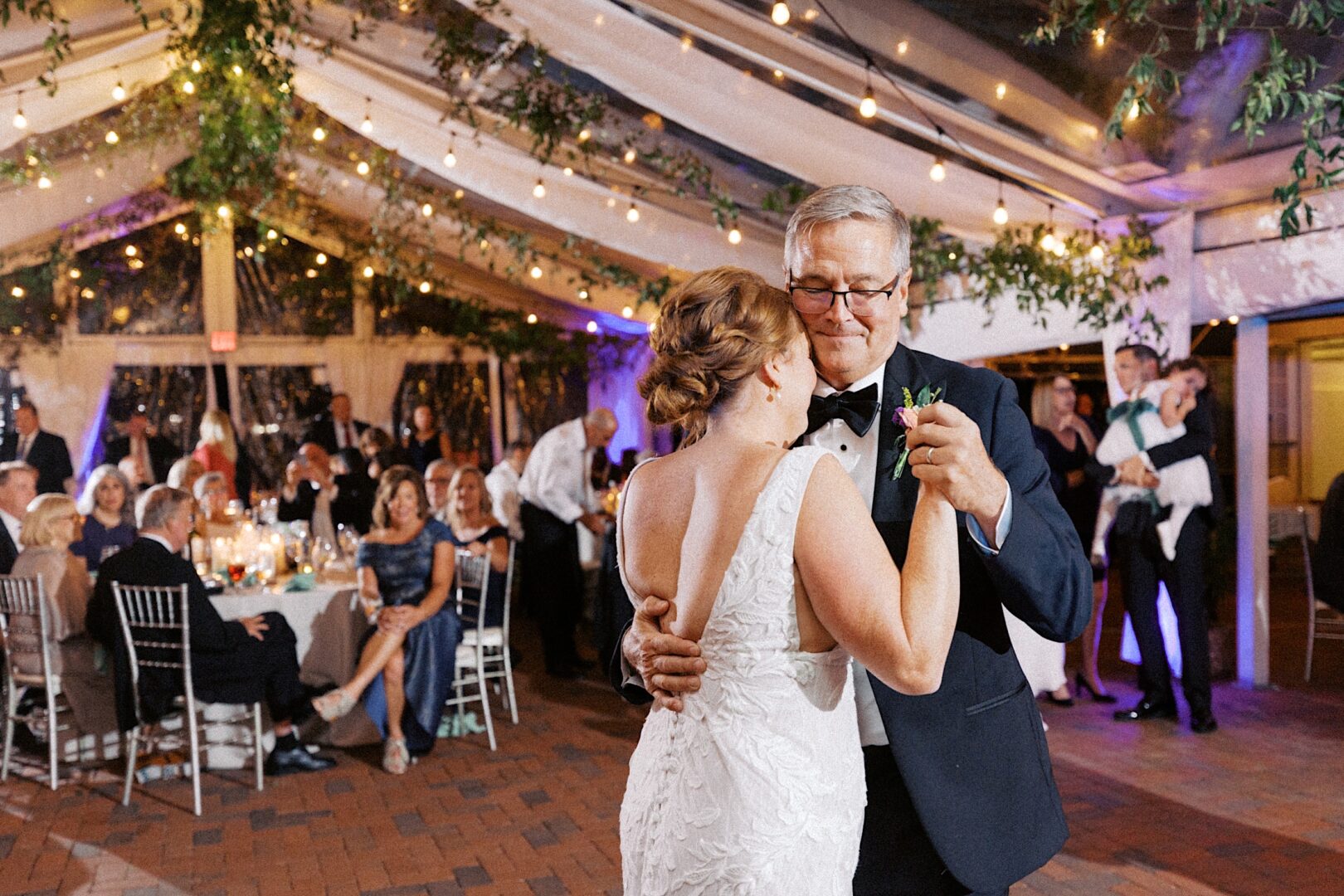 A couple dances in a warmly lit event space at the Corinthian Yacht Club of Cape May Wedding, where hanging greenery and string lights create a magical atmosphere. Guests are seated at round tables in the background, soaking in the enchantment.