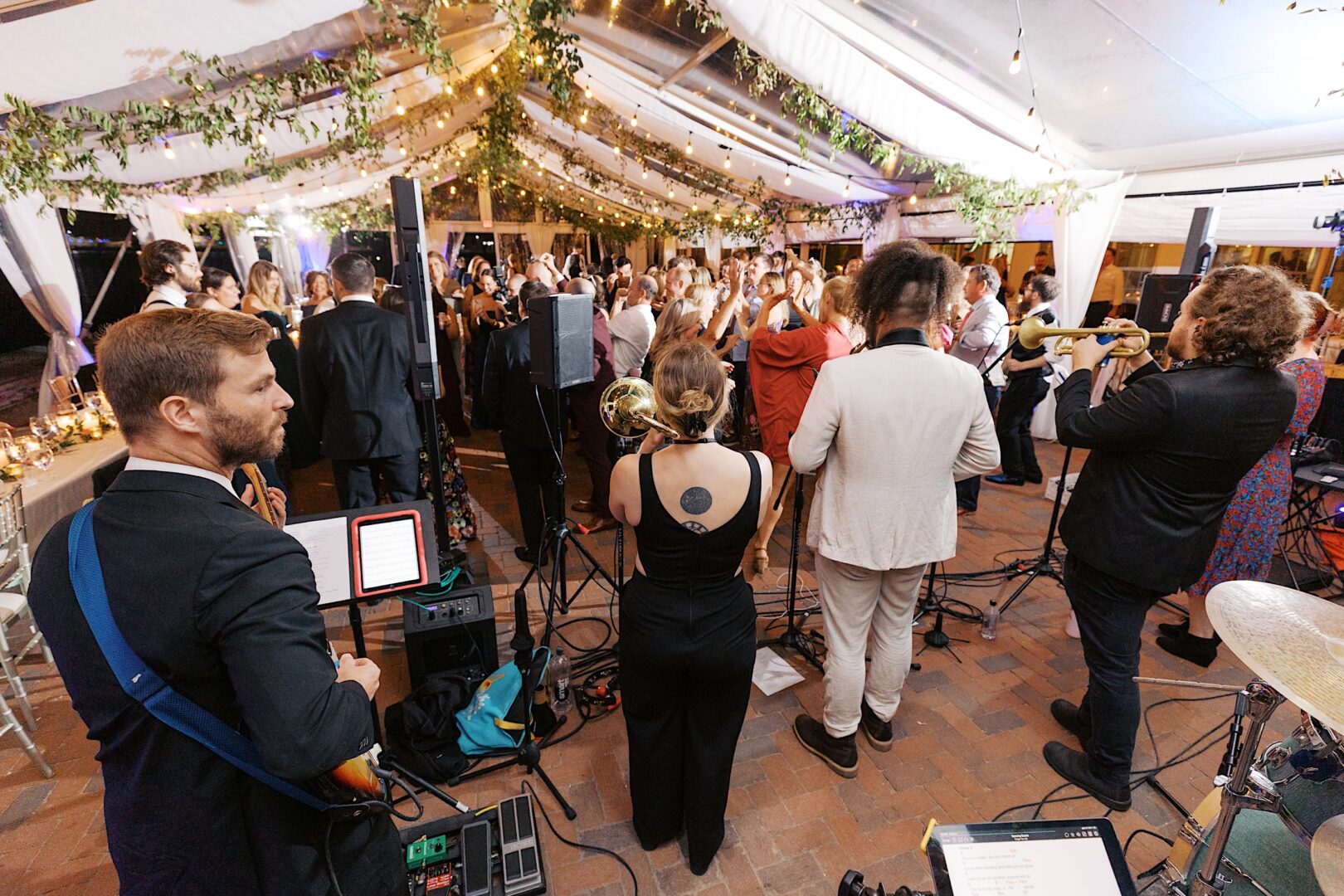 At the Corinthian Yacht Club of Cape May wedding, a band performs at an indoor event, surrounded by a lively crowd. String lights and greenery elegantly decorate the ceiling, adding charm to the celebration.