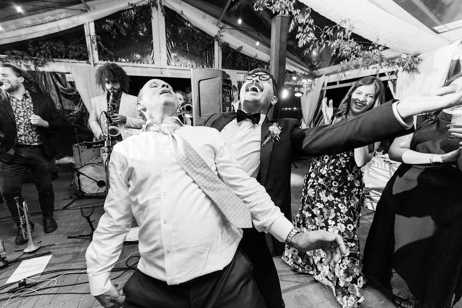 Two men joyfully dancing near musicians at an event, capturing the vibrant spirit of a Corinthian Yacht Club of Cape May Wedding. A woman in a floral dress and others add to the lively background scene.