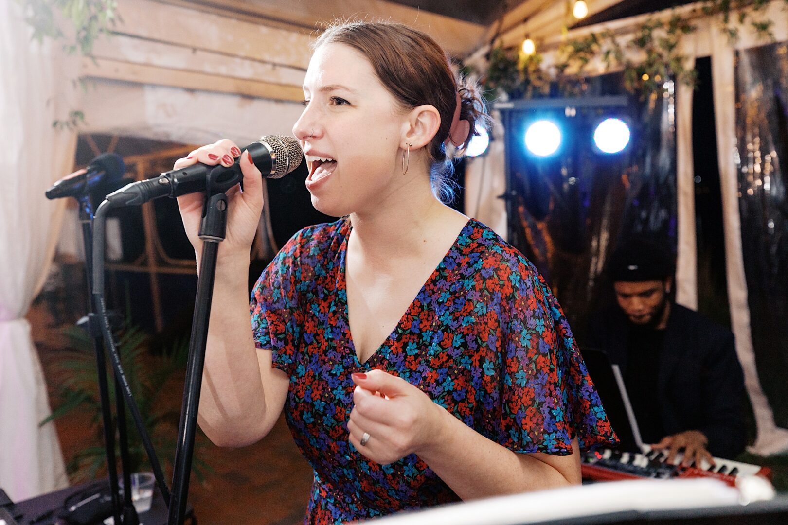 At a Corinthian Yacht Club of Cape May wedding, a woman in a floral dress passionately sings into a microphone at an indoor event, as a musician harmonizes on the keyboard in the background.