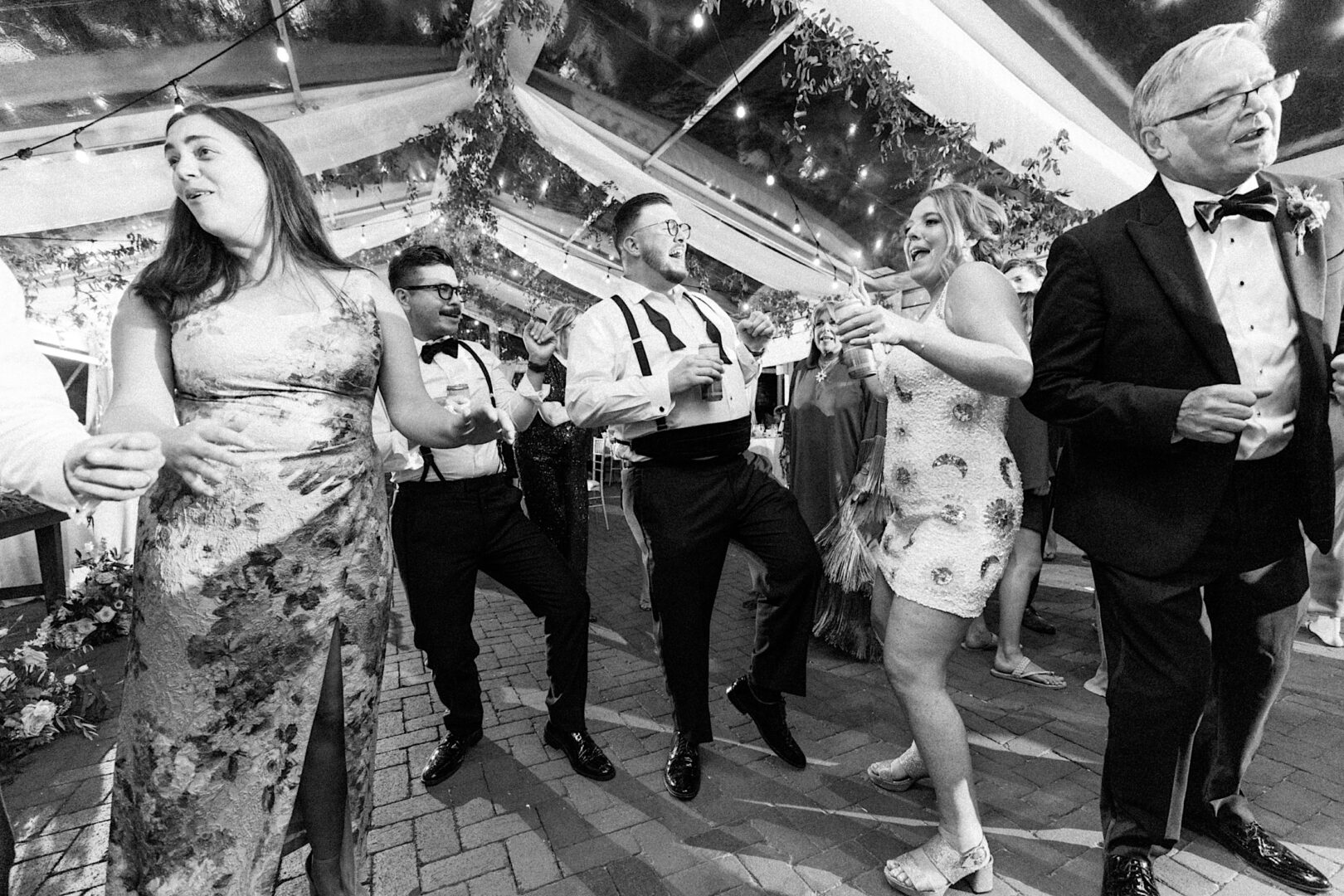 At the Corinthian Yacht Club of Cape May wedding, a group in formal attire dances joyfully under a tent adorned with romantic string lights.