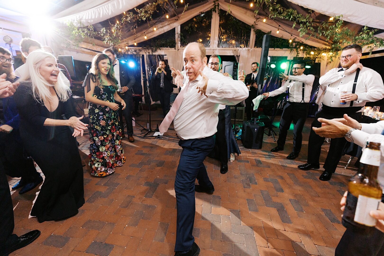 A man dances energetically at an outdoor party, surrounded by smiling people and festive lights, echoing the joyous spirit reminiscent of a Corinthian Yacht Club of Cape May wedding.