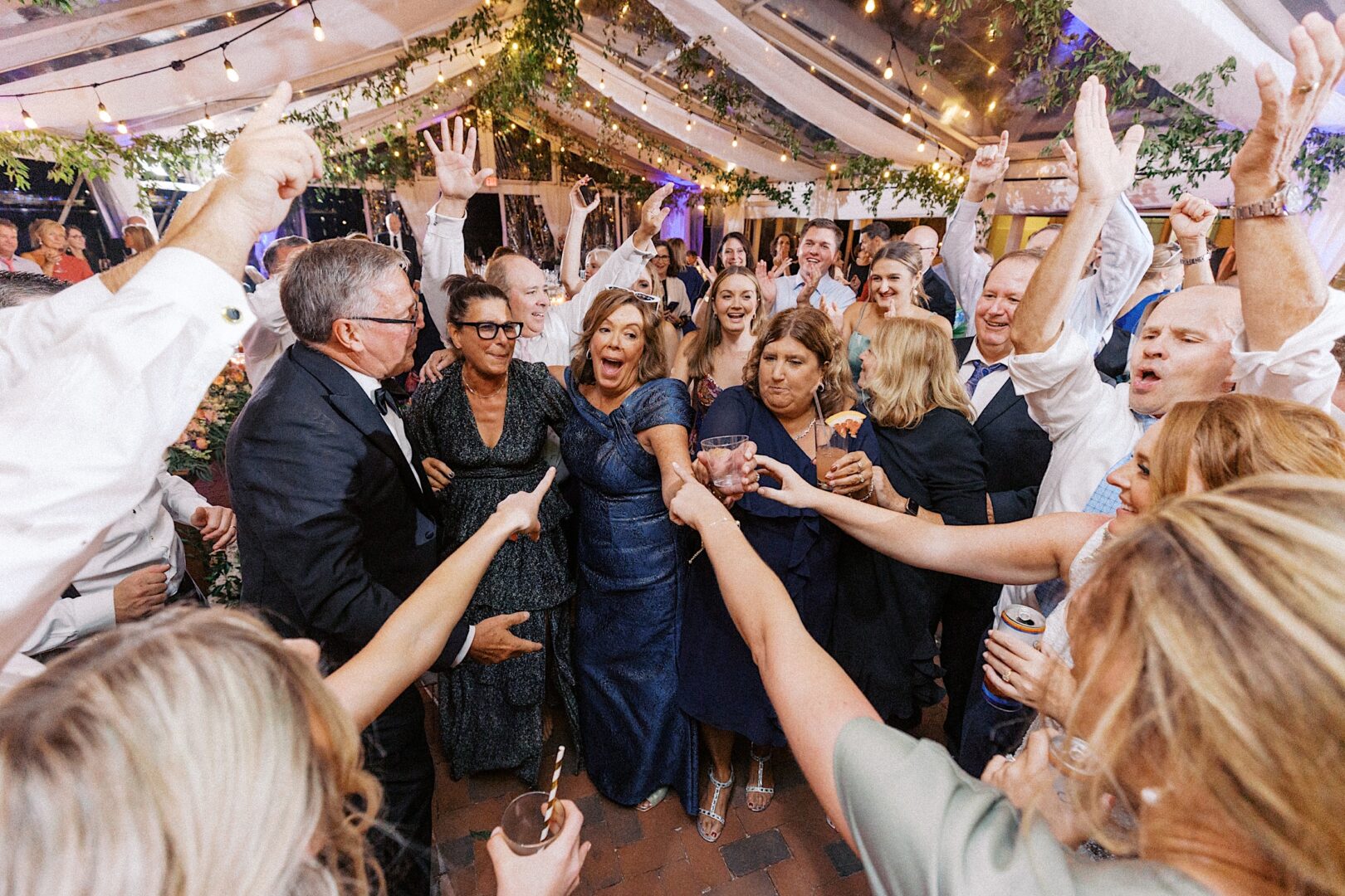 A group of people celebrating under a tent at the Corinthian Yacht Club of Cape May Wedding, adorned with string lights and greenery. Many are raising their arms and smiling, creating a joyful atmosphere.