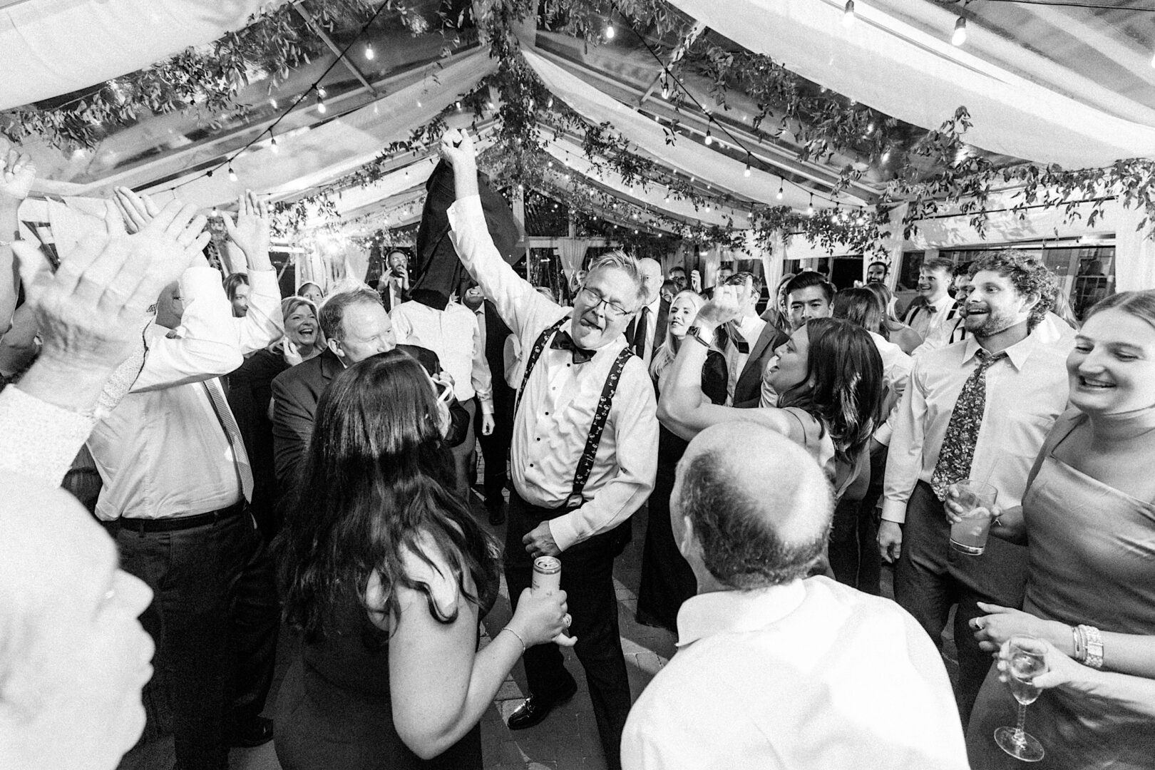 A group of people celebrating indoors under a draped ceiling with hanging greenery at the Corinthian Yacht Club of Cape May wedding, as a man in the center raises his hand, surrounded by clapping and cheering guests.