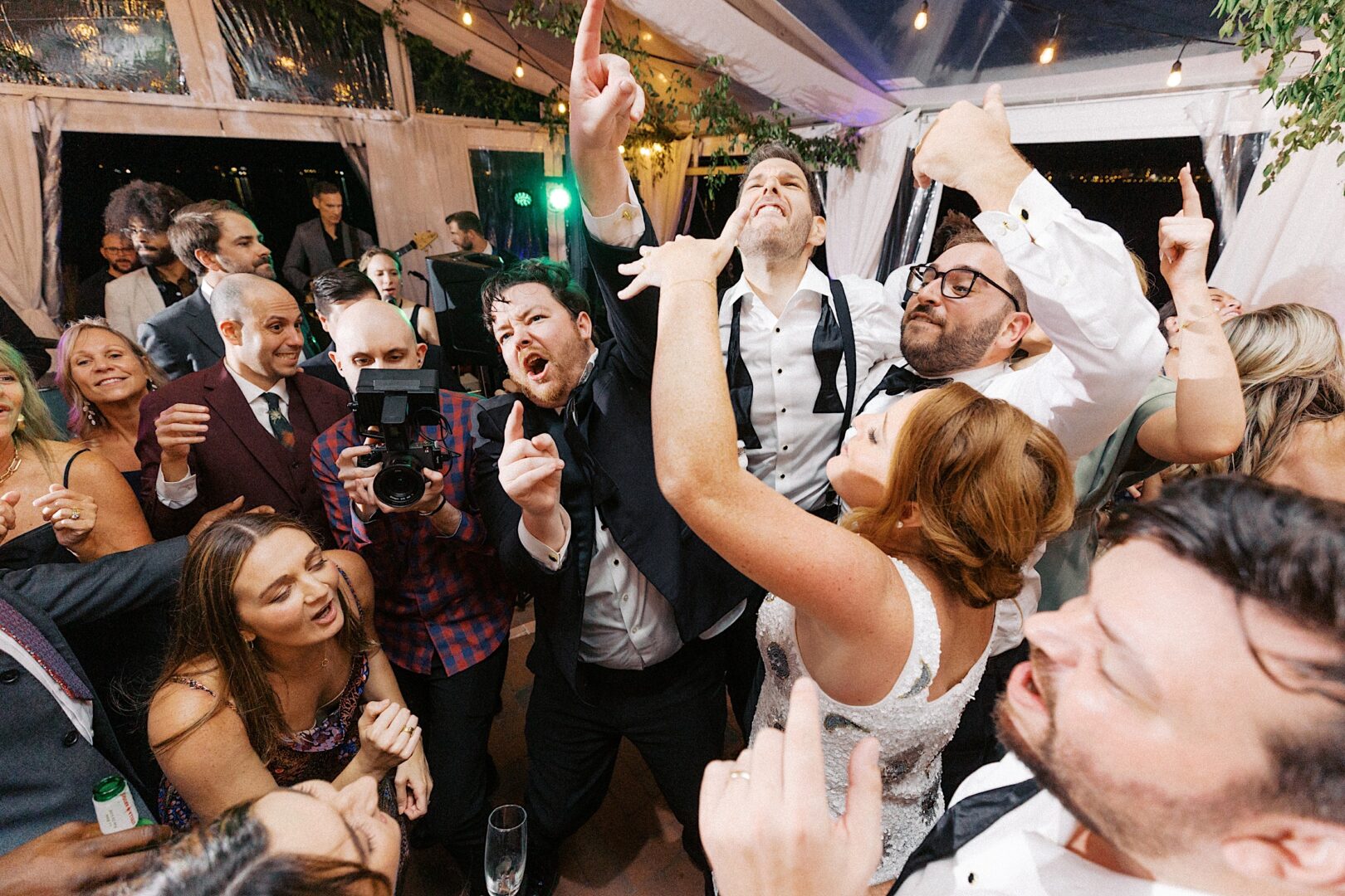 A lively group dances energetically at a party, some in formal attire, as a camera captures the joyous scene. Strings of lights hang from the ceiling, creating an enchanting atmosphere reminiscent of a Corinthian Yacht Club of Cape May wedding celebration.