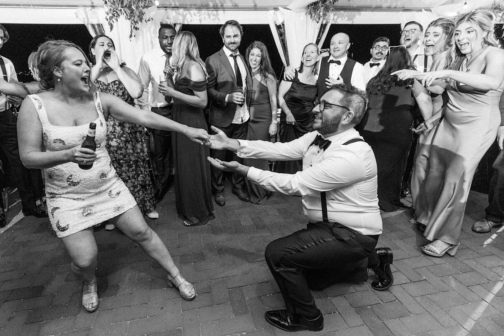 At a lively Corinthian Yacht Club of Cape May wedding, a man in formal attire kneels and reaches out to a woman holding a bottle. They are surrounded by smiling and clapping onlookers, adding to the joyous celebration.