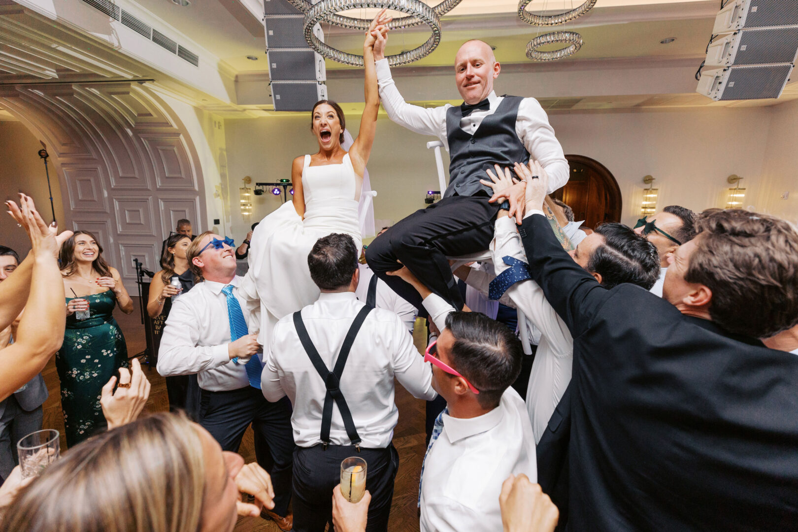 At The Tilling House wedding, a bride and groom are joyfully lifted on chairs by enthusiastic guests, while others clap and cheer in the beautifully decorated hall.