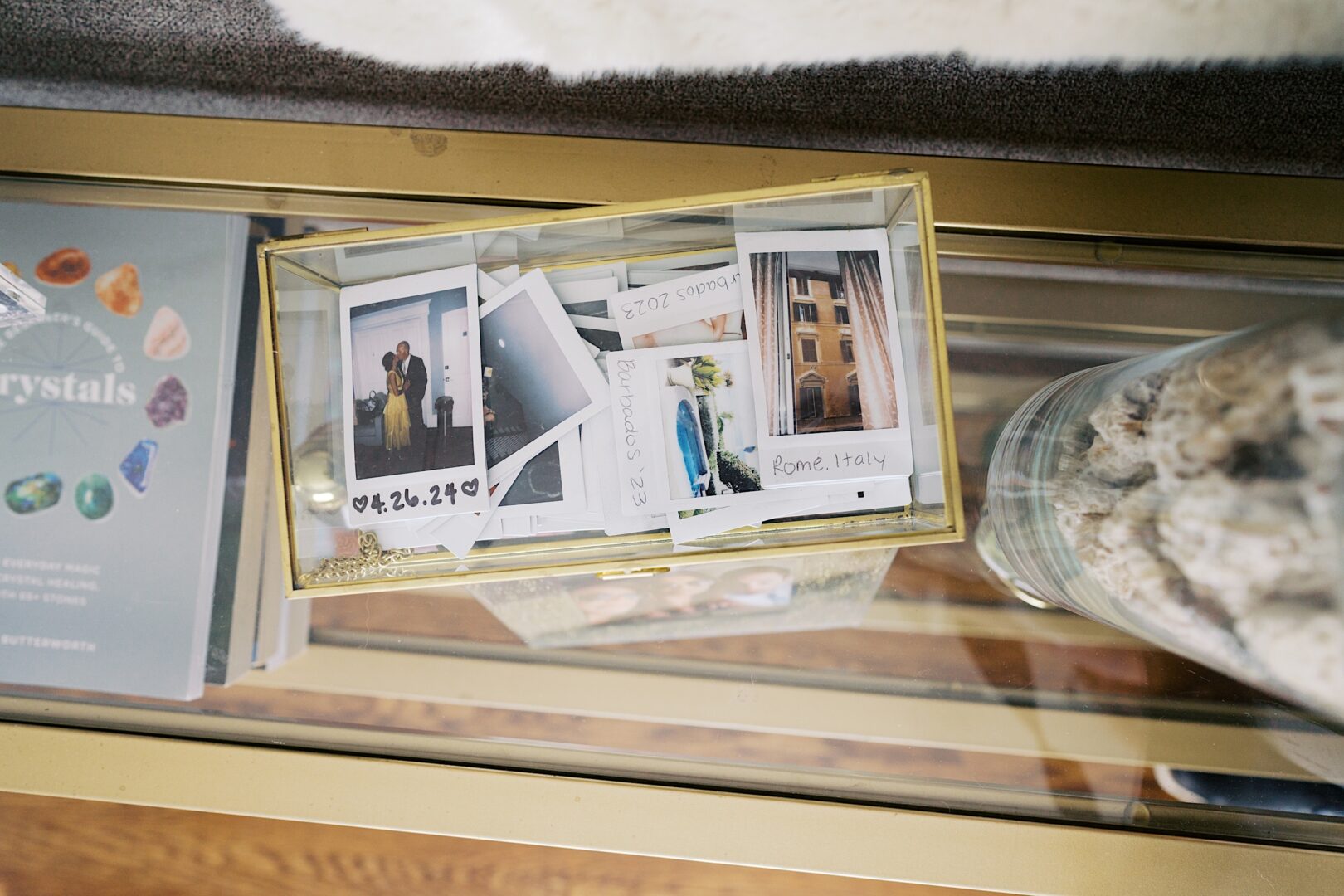 A glass container holds several Polaroid photos with handwritten notes, capturing moments from The Tilling House wedding, alongside a book titled "crystals" on a glass table.