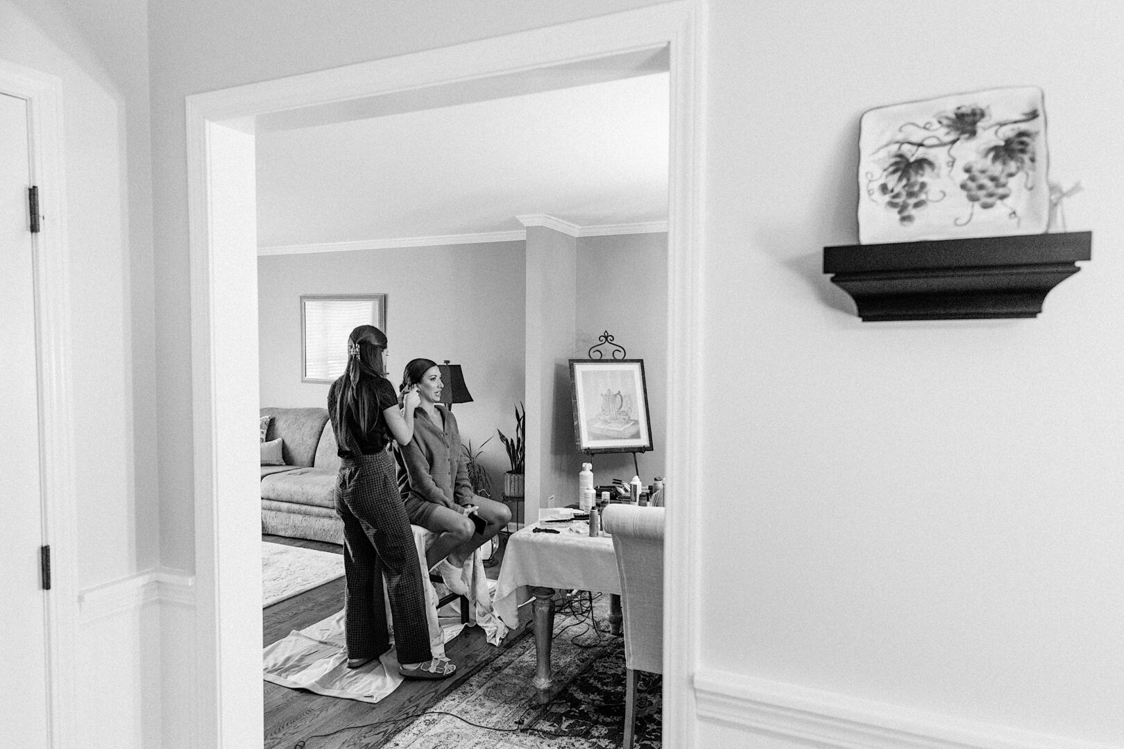 A woman is seated in a living room while another woman styles her hair for The Tilling House wedding. The room is decorated with a couch, a table filled with beauty products, and a wall shelf showcasing a decorative plate.