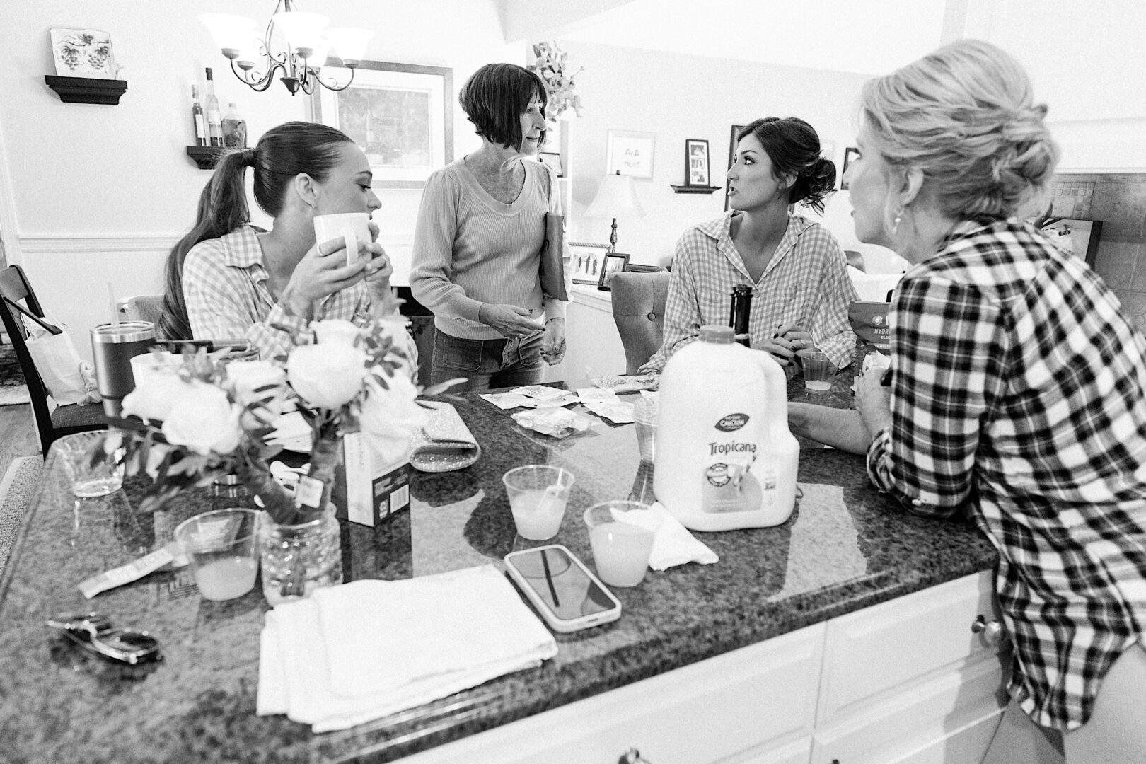 Four women gather around the kitchen island at The Tilling House wedding, chatting and holding drinks. A flower vase and juice container sit on the counter, enhancing the casual and social atmosphere.