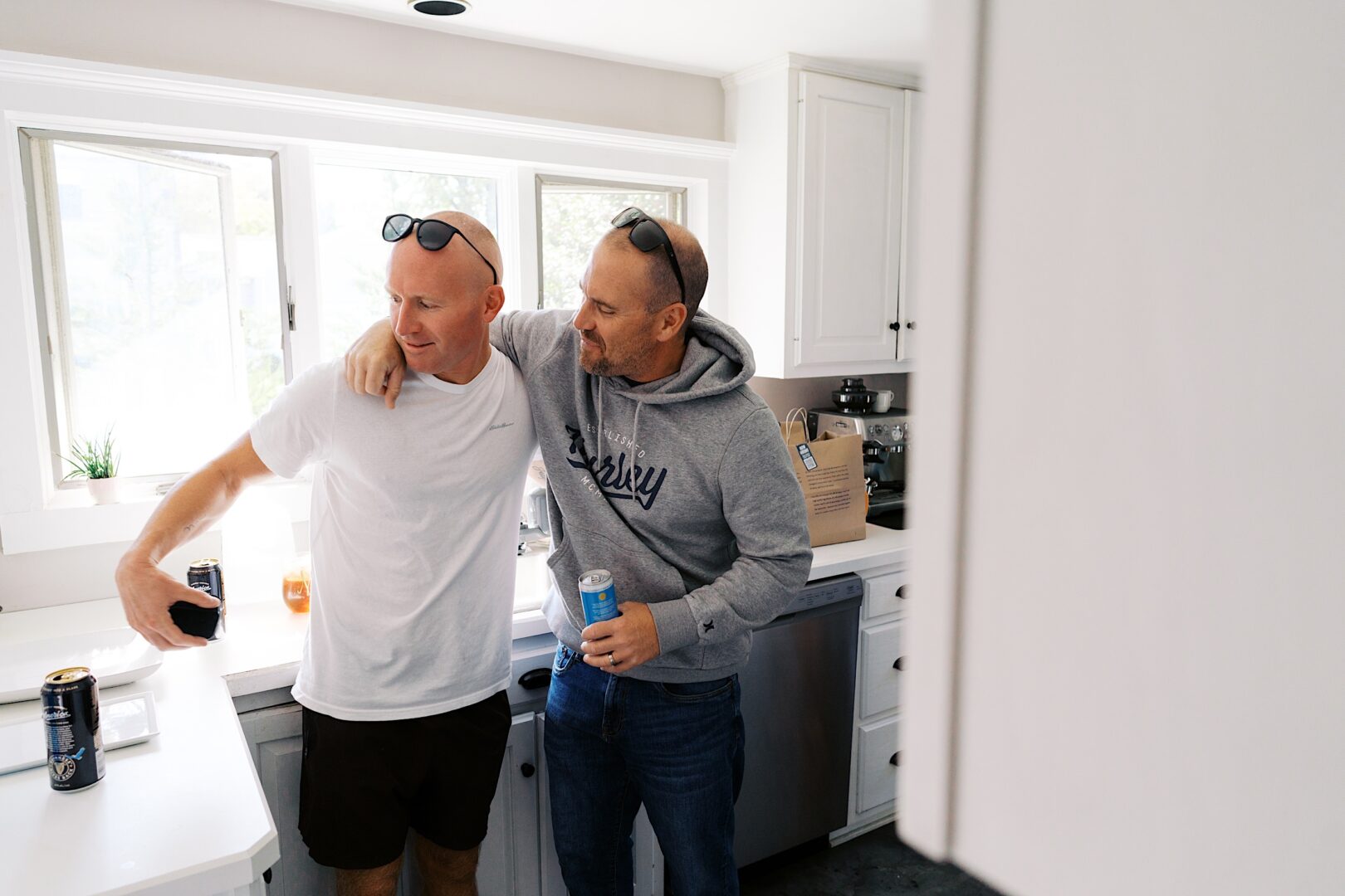 Two men stand in a kitchen; one wears a white t-shirt, the other a gray hoodie. Both have sunglasses perched on their heads and hold drinks, with one man’s arm around the other, reminiscing about The Tilling House wedding they recently attended.