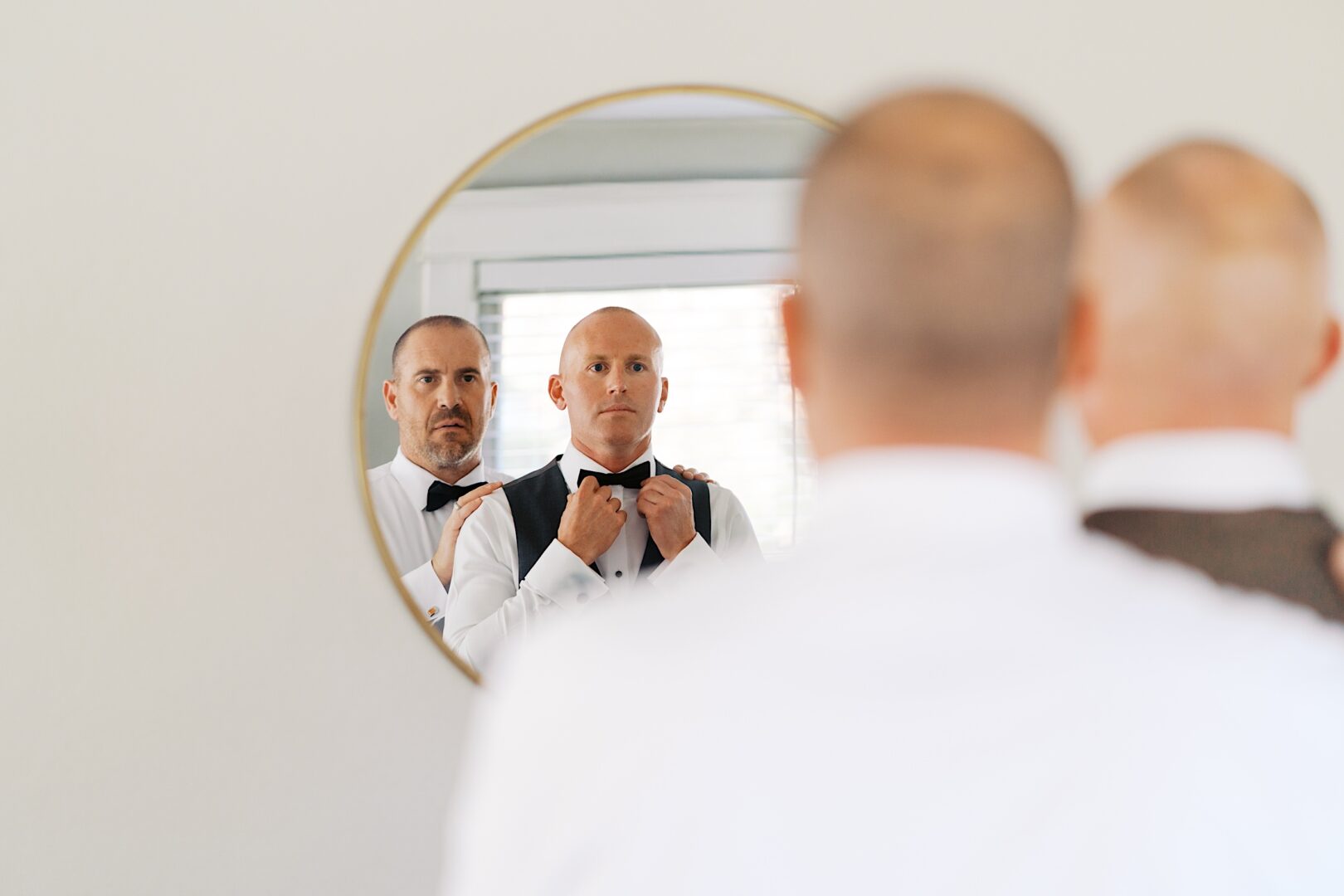 Two men are getting ready in front of a round mirror at The Tilling House wedding, adjusting their bow ties. They are dressed formally in white shirts and suit vests.