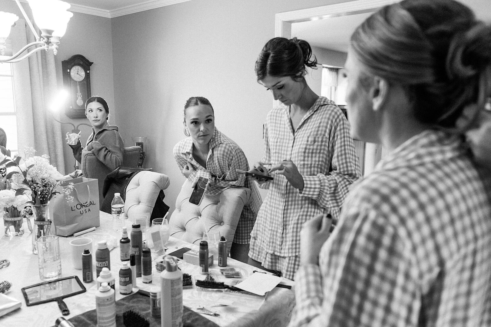 Black and white photo of four women at The Tilling House wedding, surrounded by hair and makeup products as they prepare for the event, a clock on the wall ticking away.