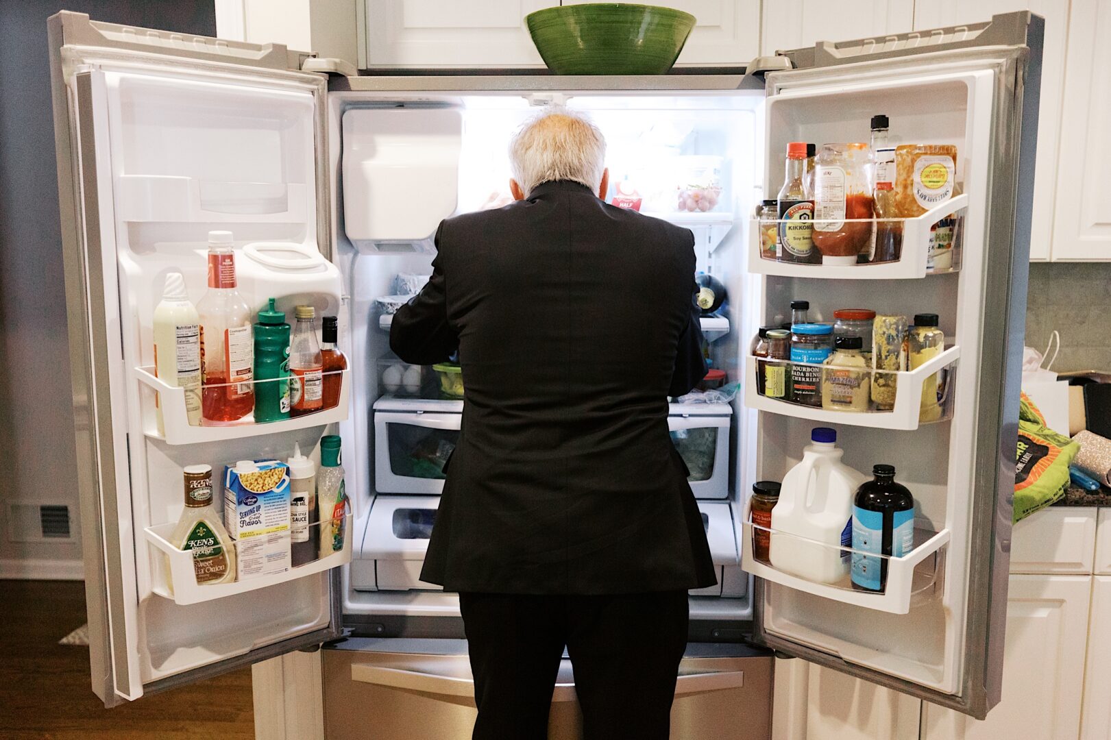 A person in a suit is standing in front of an open refrigerator, perhaps taking a break from The Tilling House wedding preparations. Inside, the fridge is filled with various food items and condiments, providing a moment of culinary contemplation amidst the festivities.