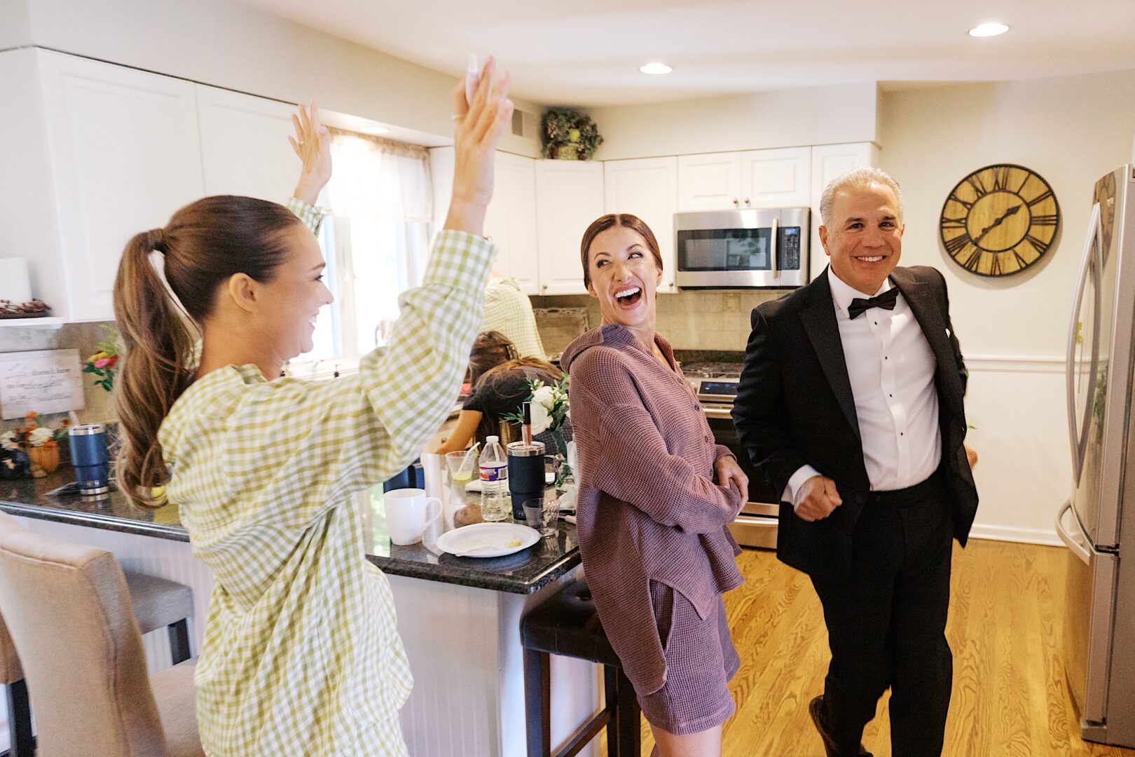 In a cozy kitchen, two women in casual attire and a man in a tuxedo share a joyful moment, reminiscent of the warmth often found at The Tilling House wedding celebrations.