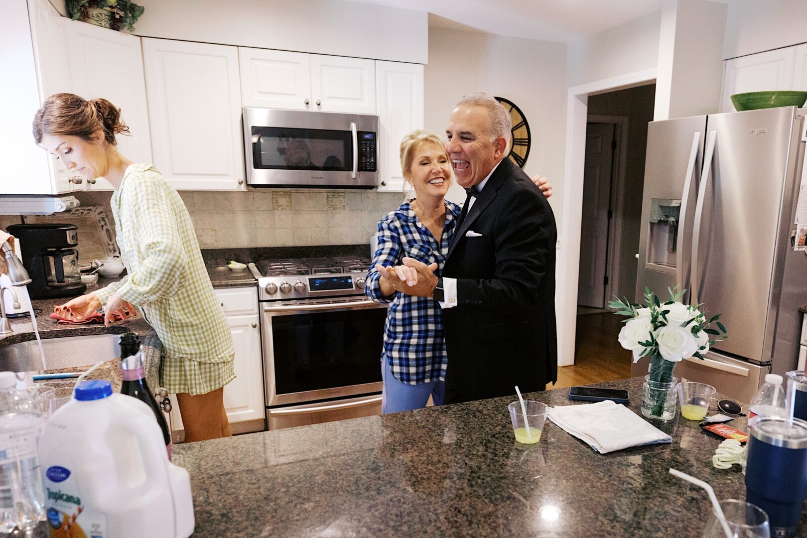 A man and woman dance in a kitchen near a counter with drinks, while another woman prepares food, reminiscent of the joyous spirit at The Tilling House wedding. Kitchen appliances are visible in the background.