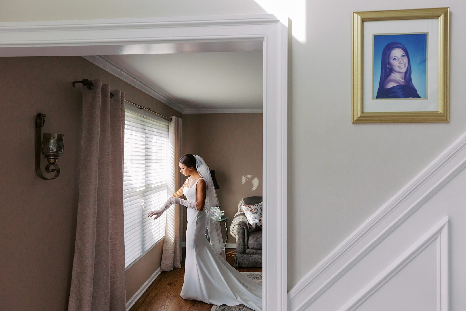 A bride in a white dress stands at a window of The Tilling House, adjusting the curtains in a sunlit room. A framed portrait hangs on the wall in the foreground.