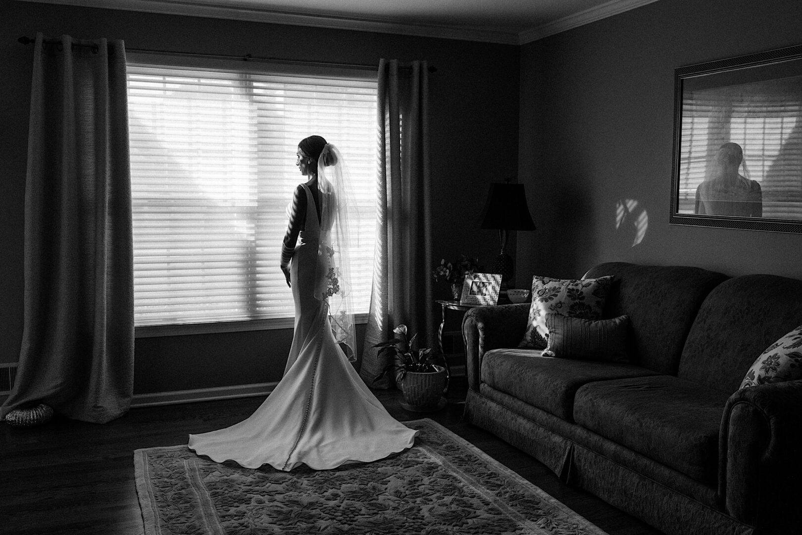 Bride in a long gown stands near a window in the dimly lit living room of The Tilling House, her reflection elegantly captured in a wall mirror.