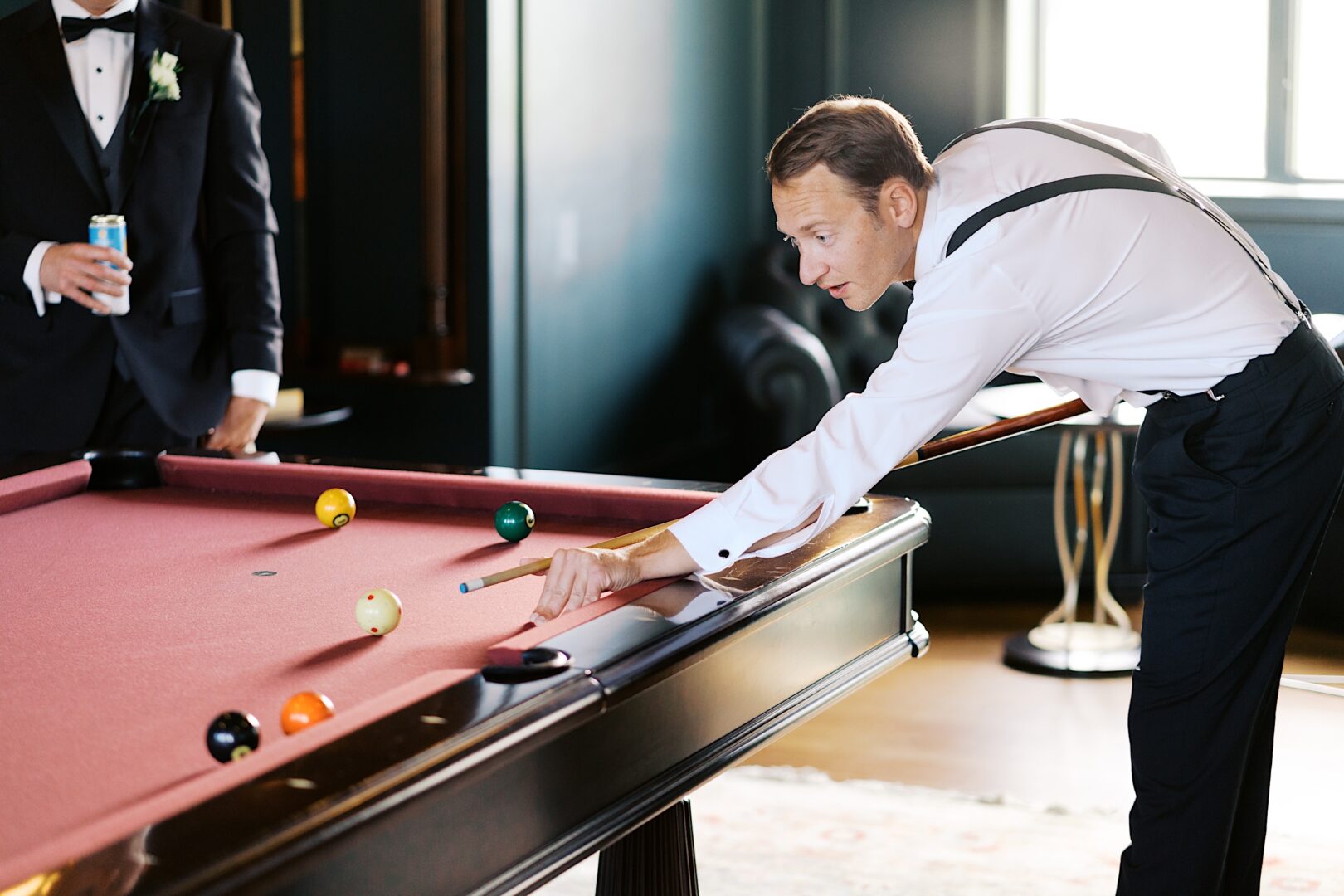 At The Tilling House wedding, a man in formal attire plays pool, expertly lining up a shot on the red table, while another man stands in the background, savoring his drink.