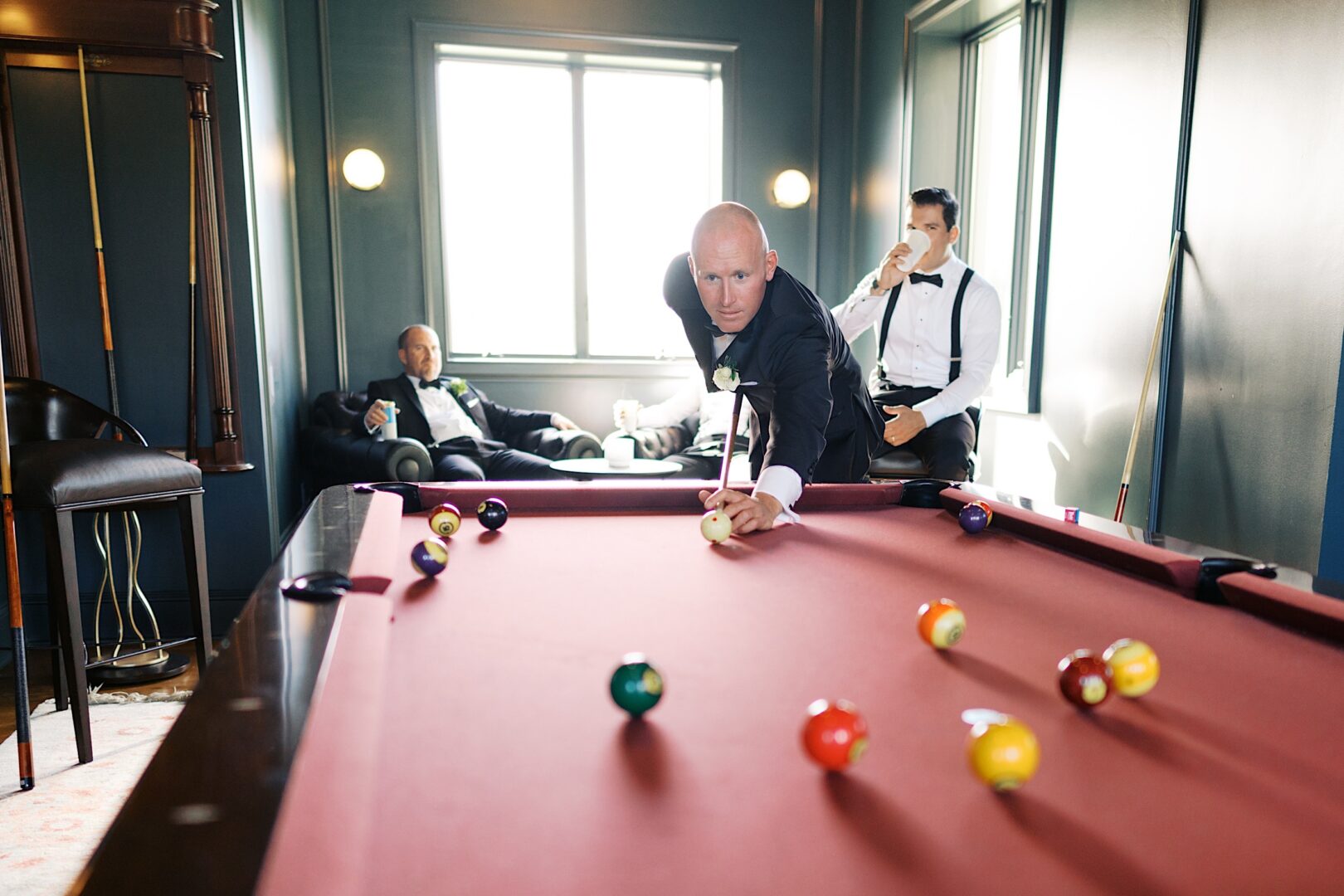 In the dimly lit room of The Tilling House wedding, a man in formal attire skillfully plays pool while two other men watch from a nearby couch.