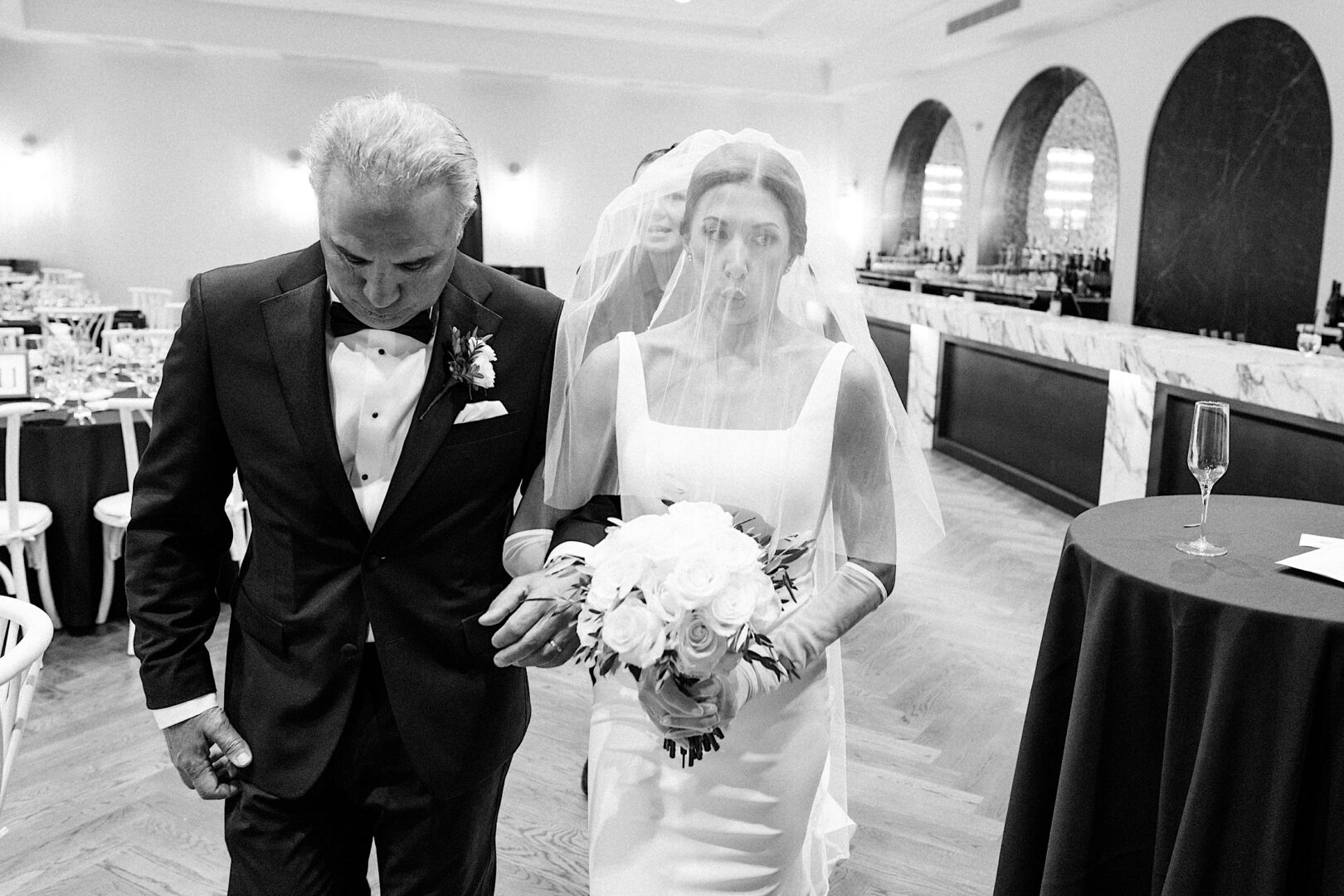 A bride holding a bouquet walks alongside a man in a tuxedo at The Tilling House wedding. They are indoors near a bar with arched mirrors, evoking an elegant and formal atmosphere for this special event.