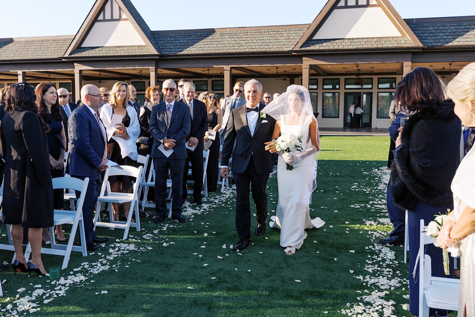 At The Tilling House wedding, a bride gracefully walks down an outdoor aisle with an older man, surrounded by guests seated on white chairs amidst scattered flower petals on the grass.