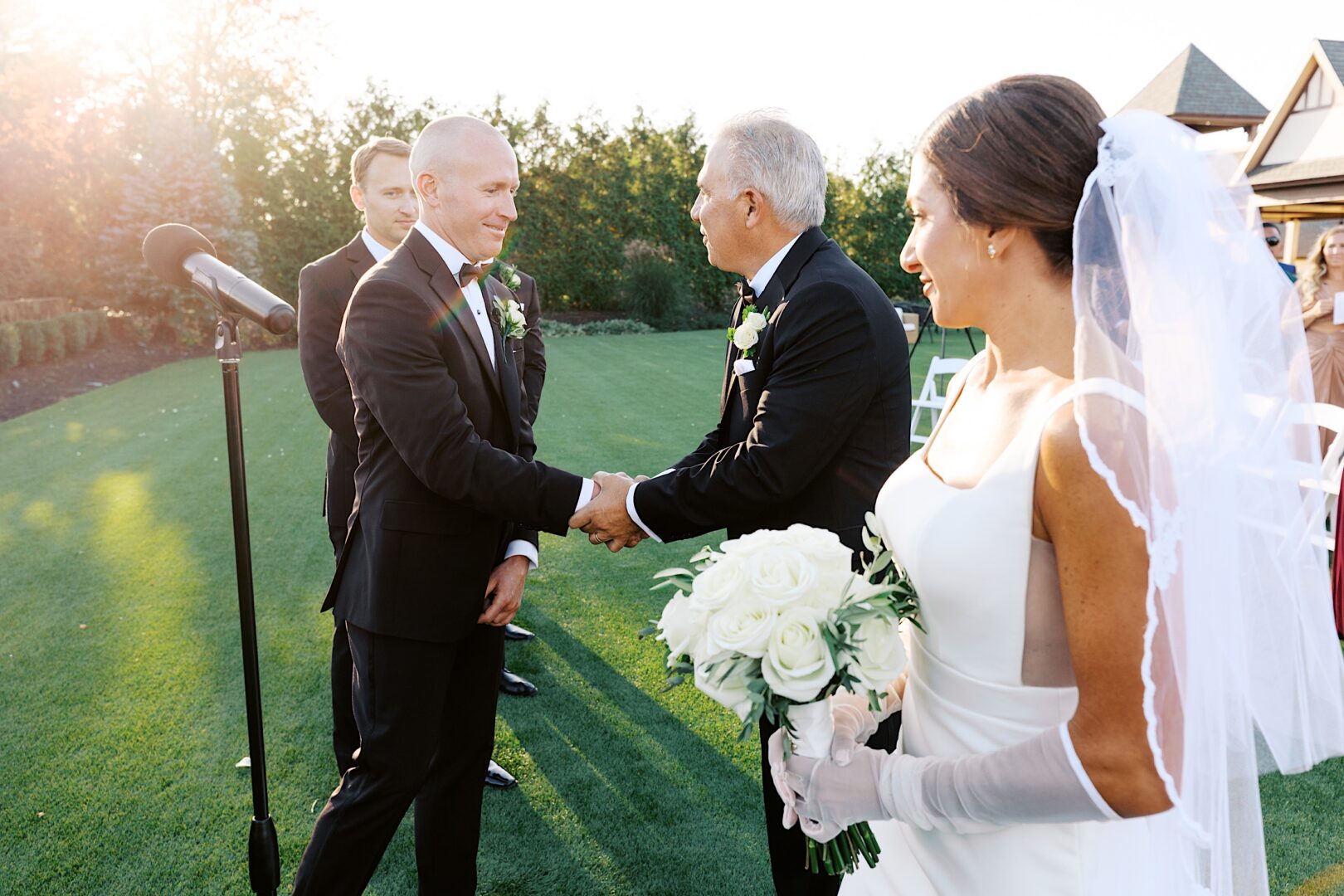 At The Tilling House wedding, a smiling bride with a bouquet looks on as the groom shakes hands with an older man during the outdoor ceremony.