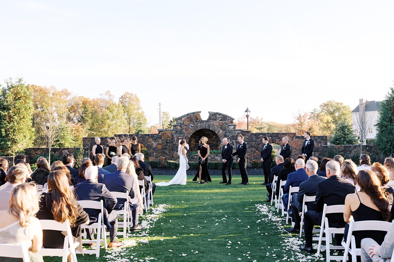 An outdoor wedding ceremony at The Tilling House, with guests seated on both sides facing the couple and officiant in front of a stone wall. Bridesmaids and groomsmen stand elegantly on either side.