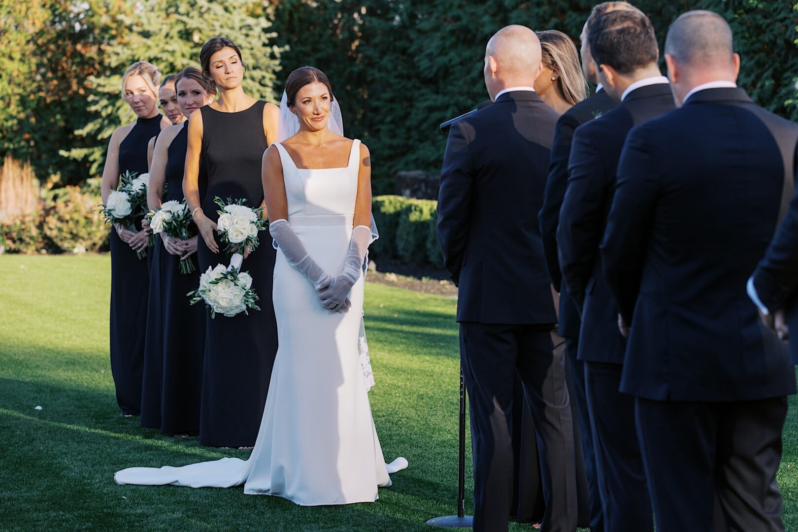 During The Tilling House wedding, a bride in a white dress and gloves stands with bridesmaids in black dresses, facing the groomsmen in suits amidst an enchanting outdoor ceremony.