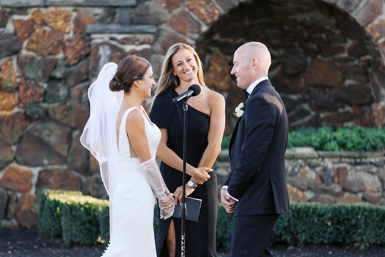 The bride and groom stand facing each other during The Tilling House wedding ceremony, with a smiling officiant between them against a backdrop of elegant stone architecture.