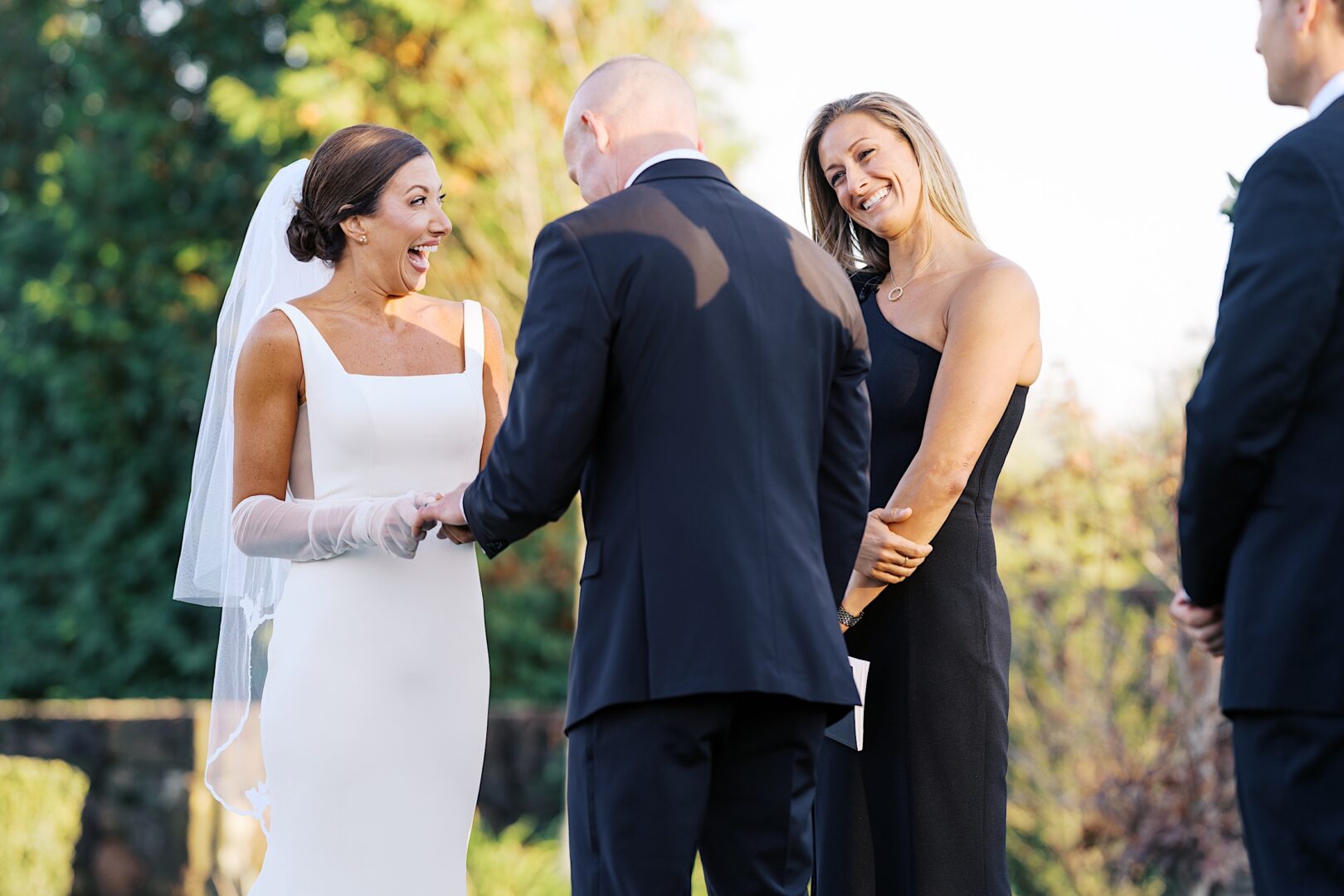 During The Tilling House wedding, a bride and groom exchange rings amidst the picturesque outdoor setting. A woman in a black dress stands beside them, smiling warmly.