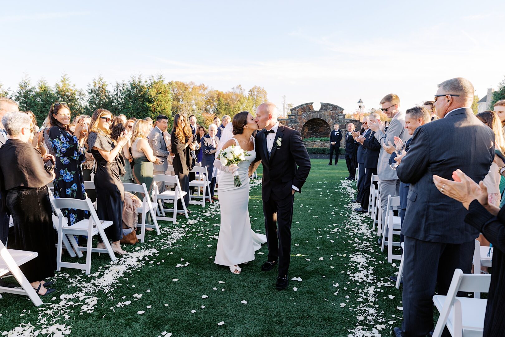 At The Tilling House wedding, a newlywed couple kisses while walking down an outdoor aisle, surrounded by clapping guests on both sides.