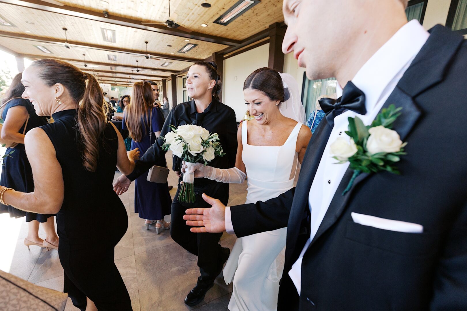 At The Tilling House wedding, a bride in a white dress and gloves walks with her groom in a black suit. She holds a bouquet of white roses, surrounded by guests in formal attire at this elegant indoor event.